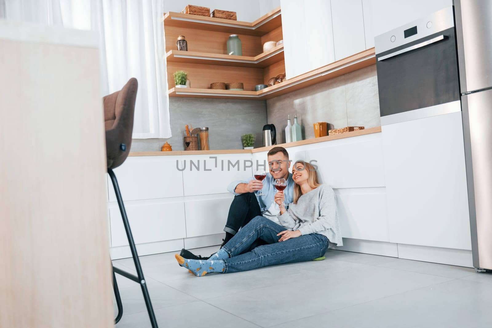 Drinking wine. Couple sitting on the floor of modern kitchen by Standret