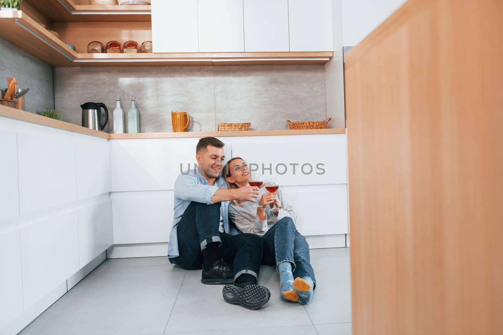 Drinking wine. Couple sitting on the floor of modern kitchen by Standret