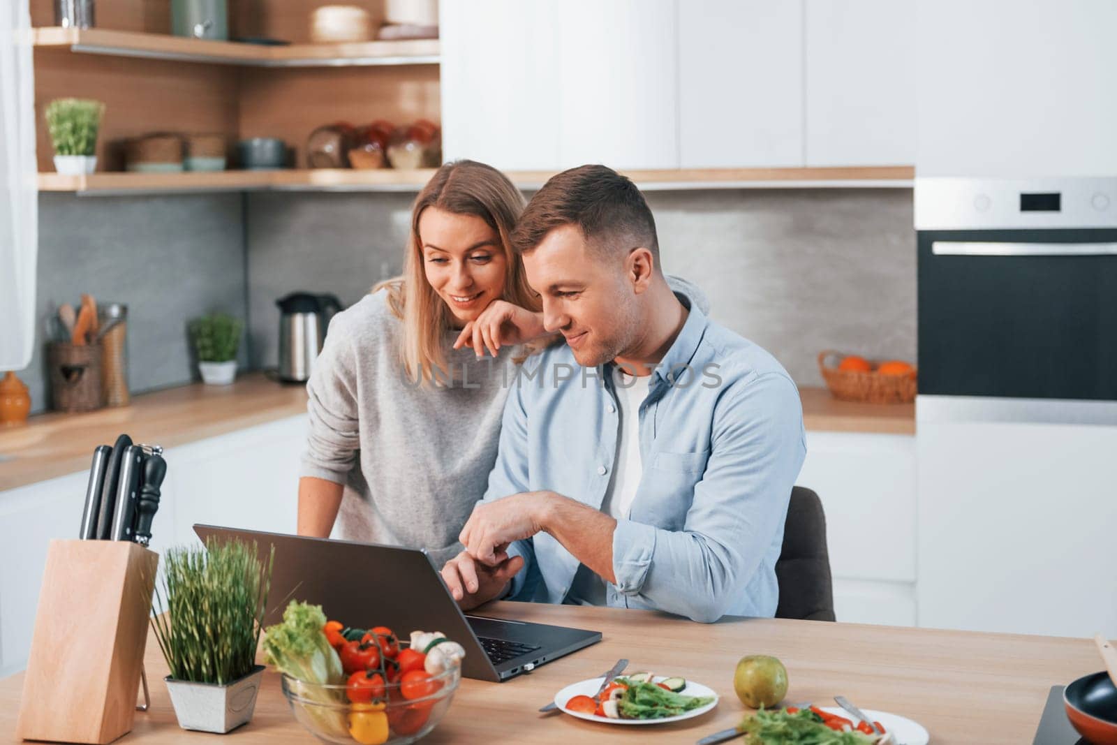 Looking at laptop. Couple preparing food at home on the modern kitchen.