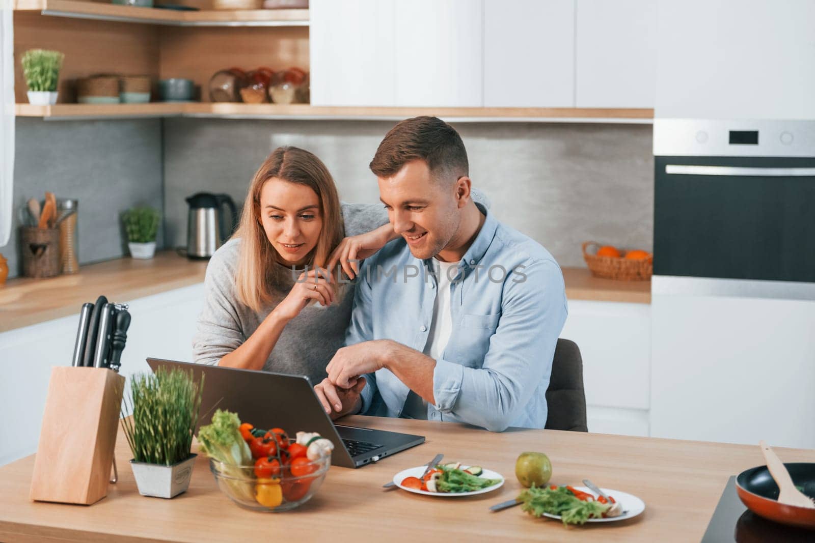 Laptop is on the table. Couple preparing food at home on the modern kitchen.