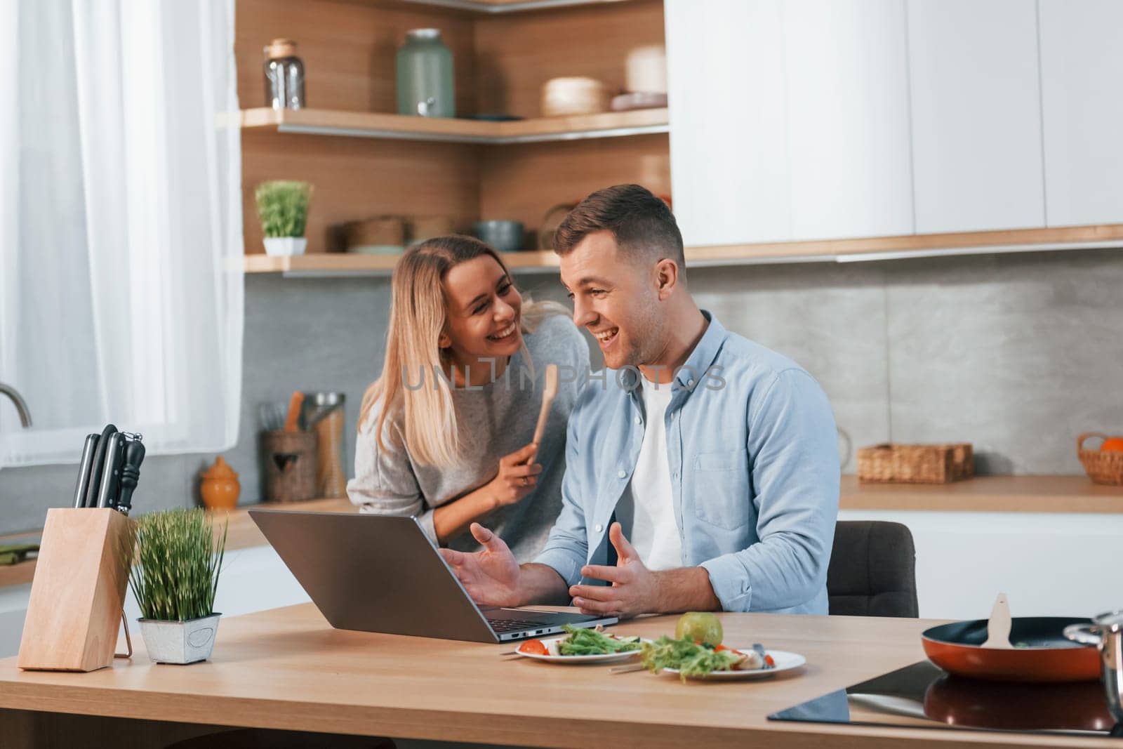 Modern laptop is on the table. Couple preparing food at home on the modern kitchen.