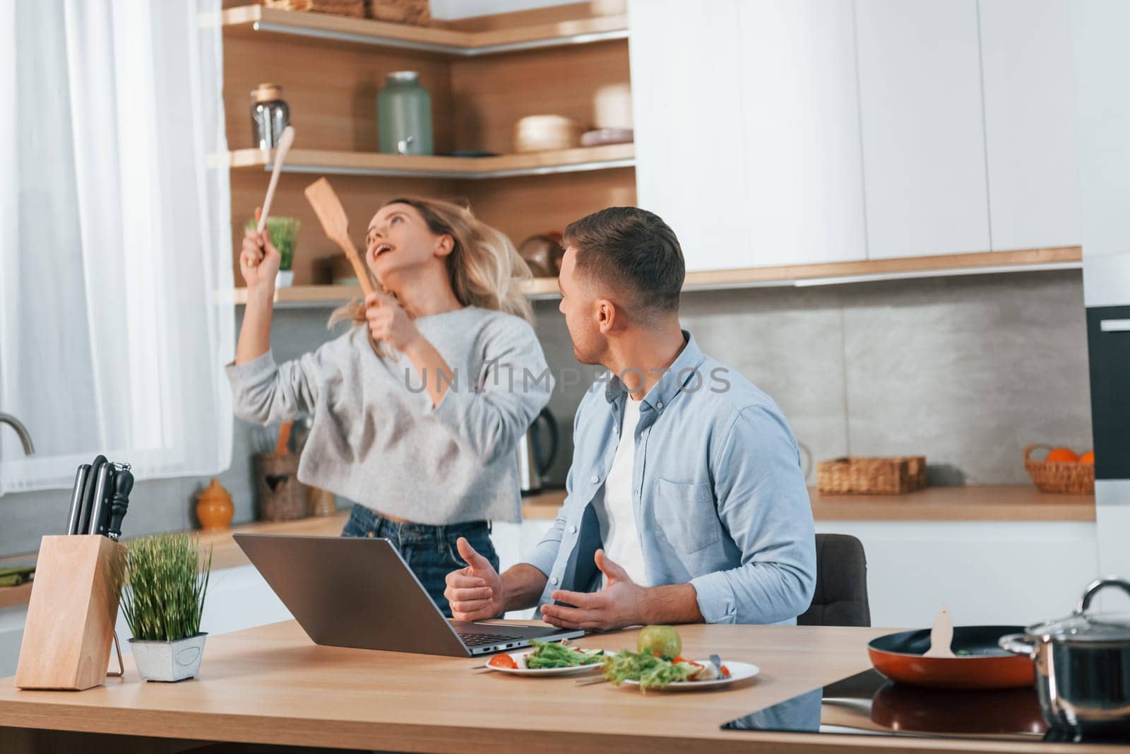 Modern laptop is on the table. Couple preparing food at home on the modern kitchen.