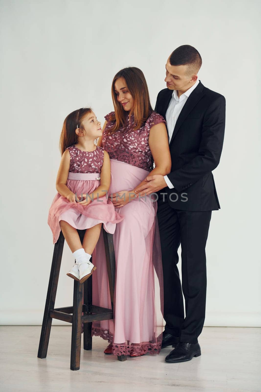 Girl is sitting on the chair. Happy couple with little daughter is standing in the studio.