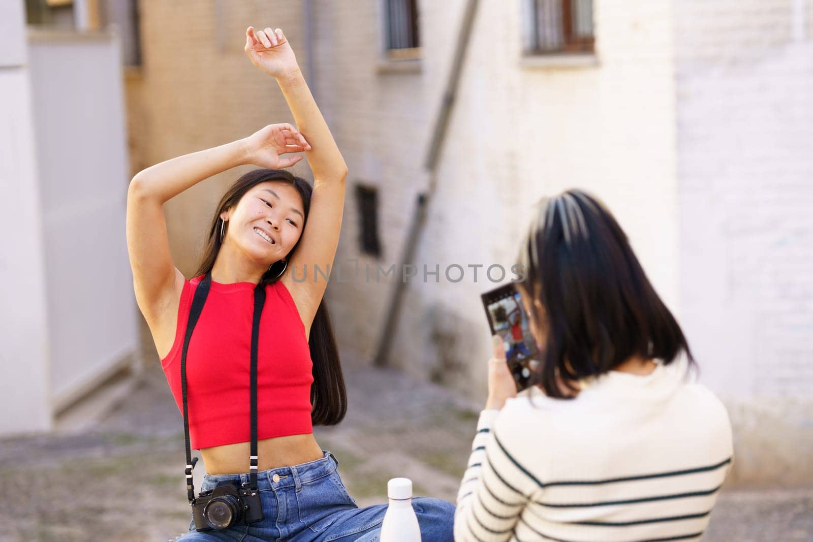 Happy young Asian female tourist in casual clothes posing for photo with raised arms in front of girlfriend against brick building in old town