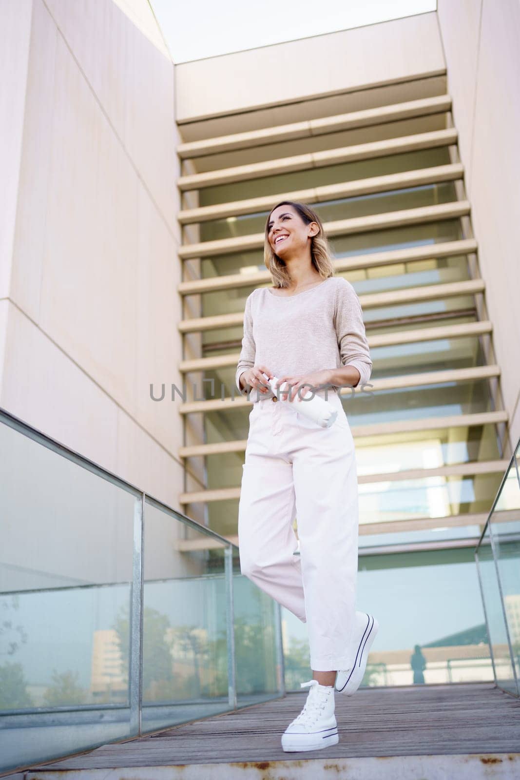 Low angle of glad stylish woman with zero waste bottle of water smiling and looking away while walking on bridge outside contemporary building