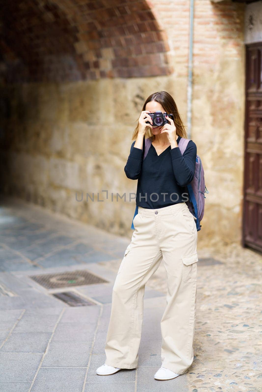 Full body of young female tourist in casual outfit with backpac,standing near aged brick building with arched passage and taking pictures on photo camera during sightseeing trip