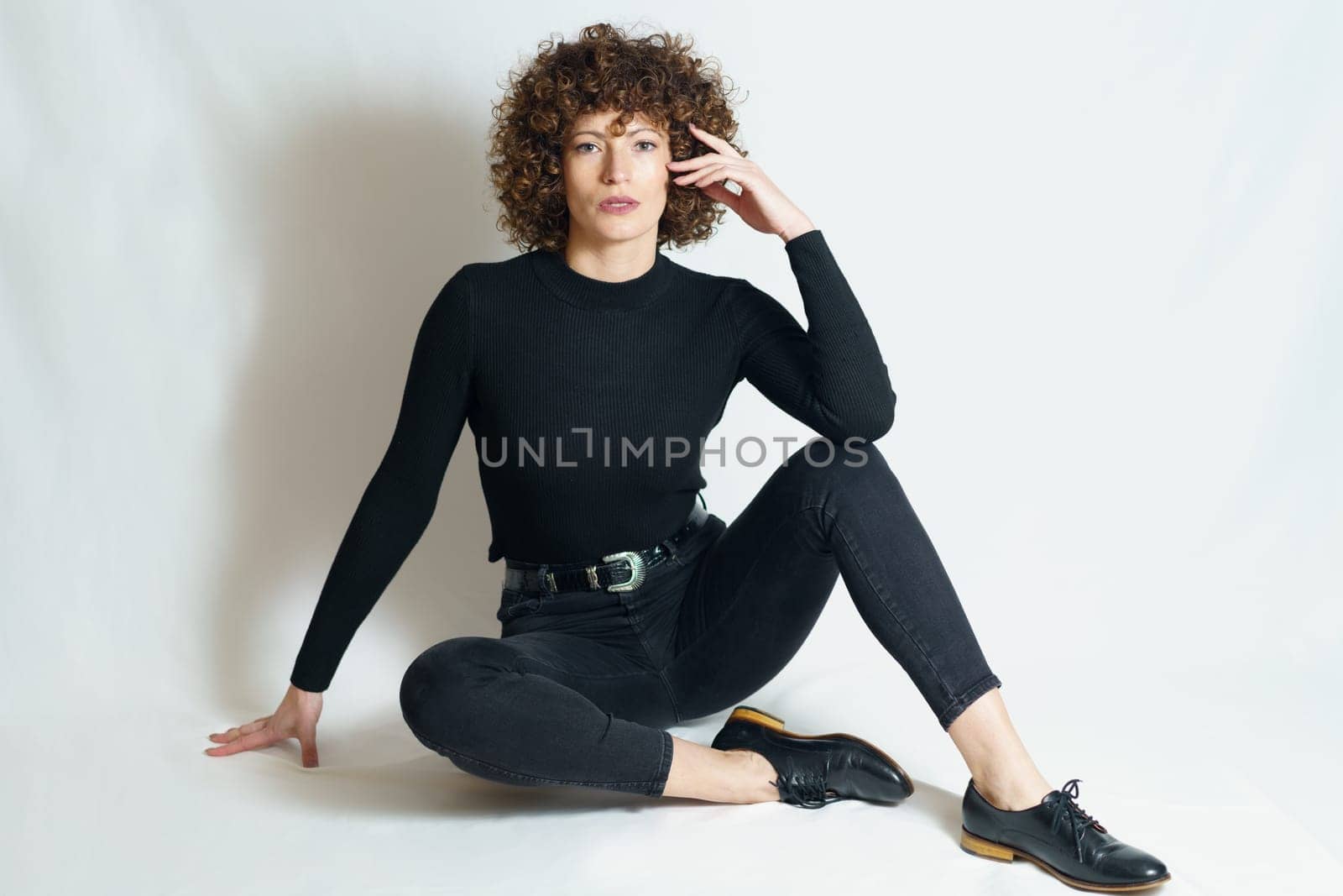 Full body of young female model, in trendy outfit relaxing with support of hand on floor and touching cheek with fingers while leaning on raised leg and looking at camera.