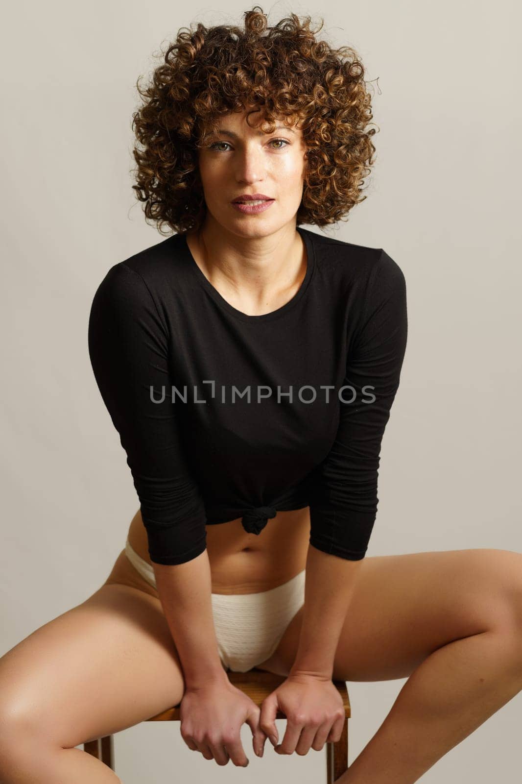 Smiling young female model, with curly hair relaxing on wooden stool with hands near seat and legs placed on steps while looking at camera against gray background