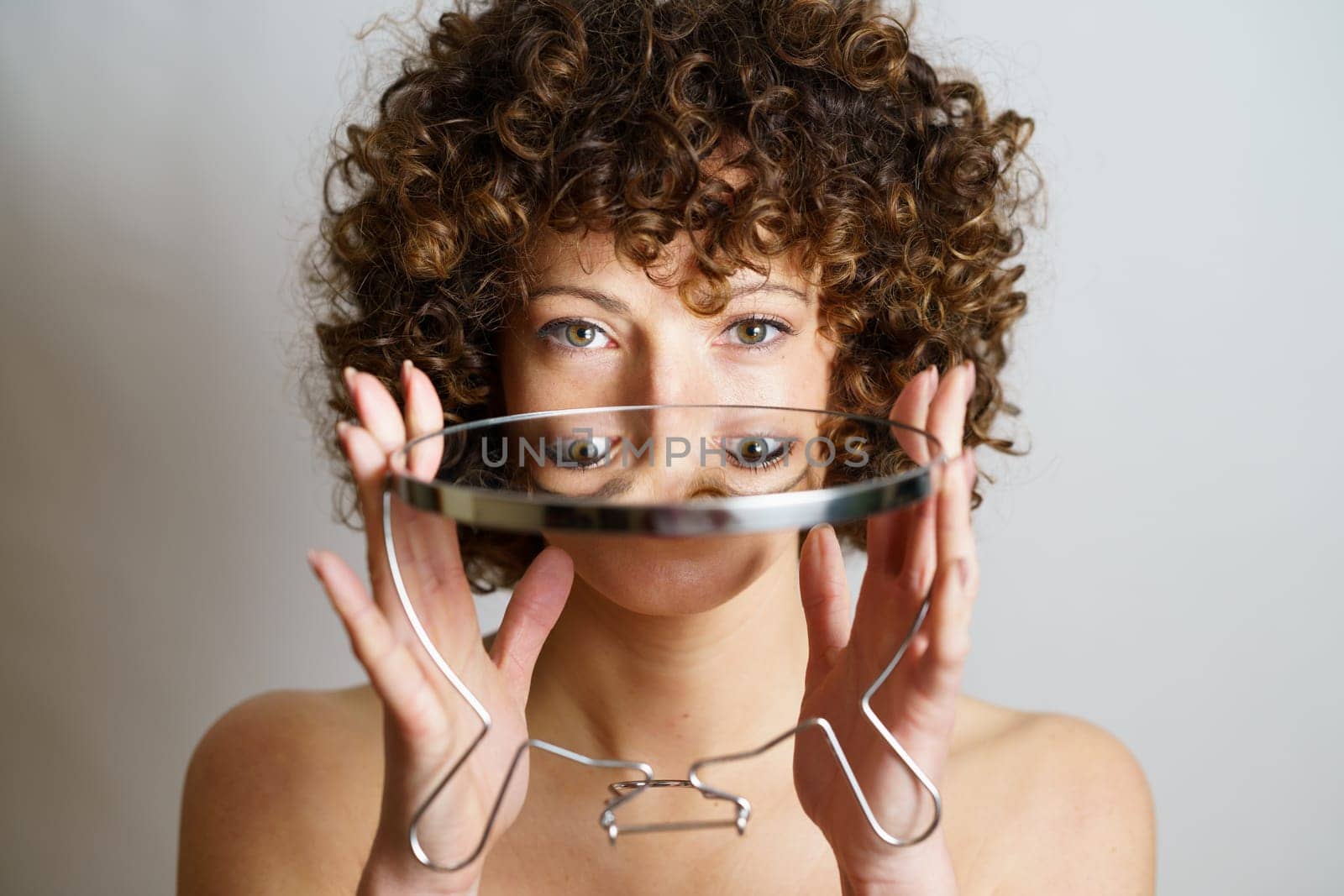 Attractive young curly haired female model holding round mirror in hands with reflection of face and eyes while looking at camera against gray background