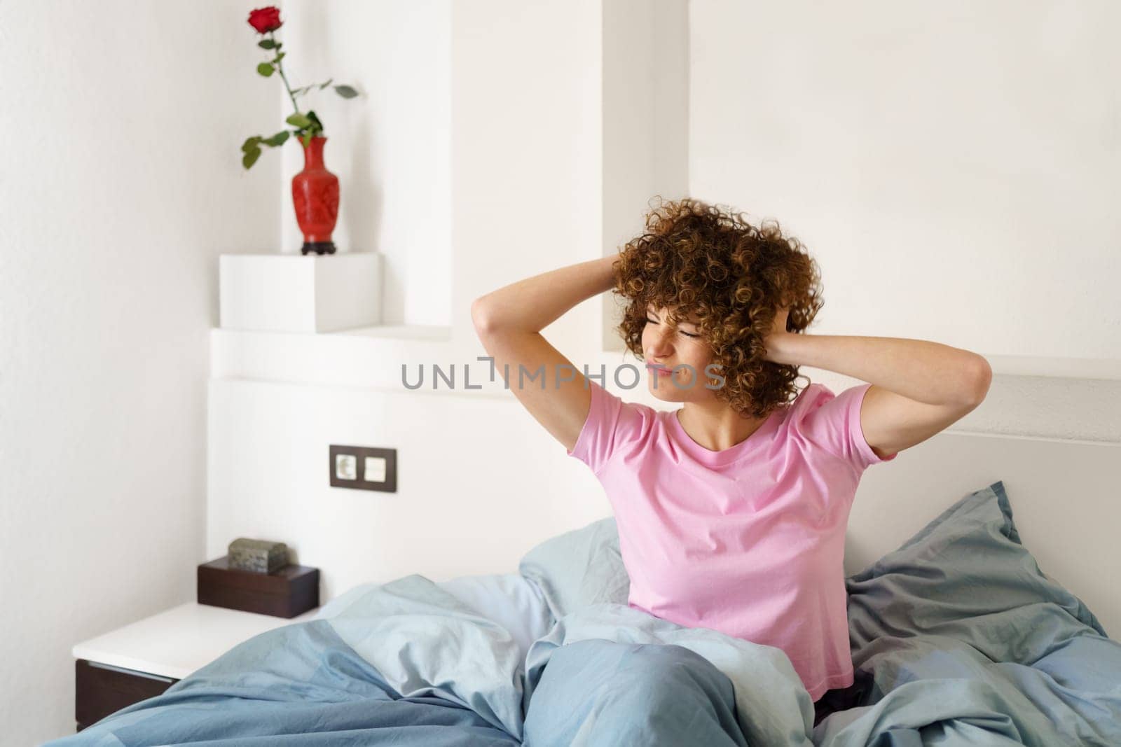 Curly haired female touching head and looking away while sitting on bed and getting up in morning