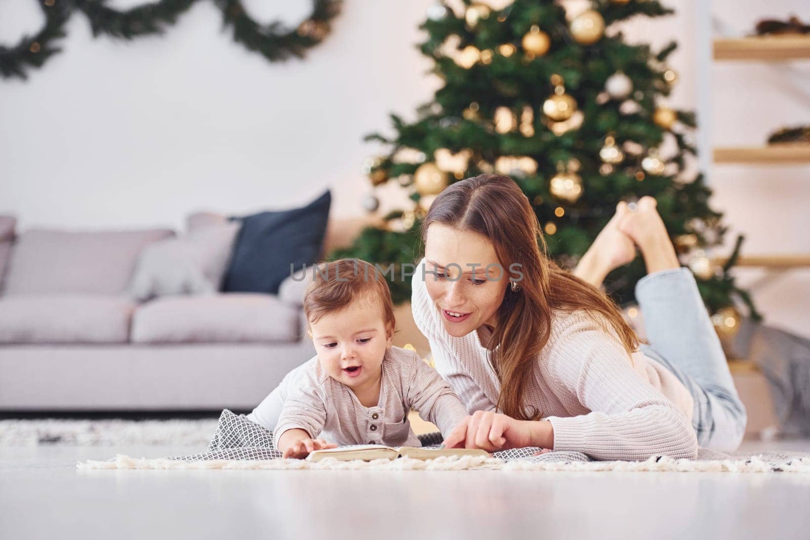 With book that is on the ground. Mother with her little daughter is indoors at home together.