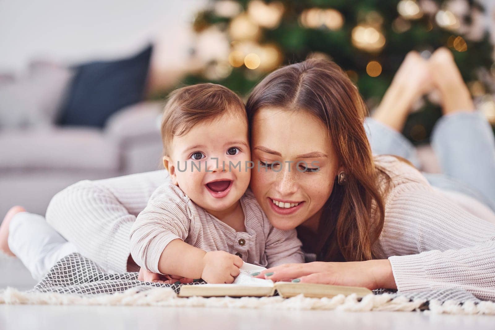 With book that is on the ground. Mother with her little daughter is indoors at home together.