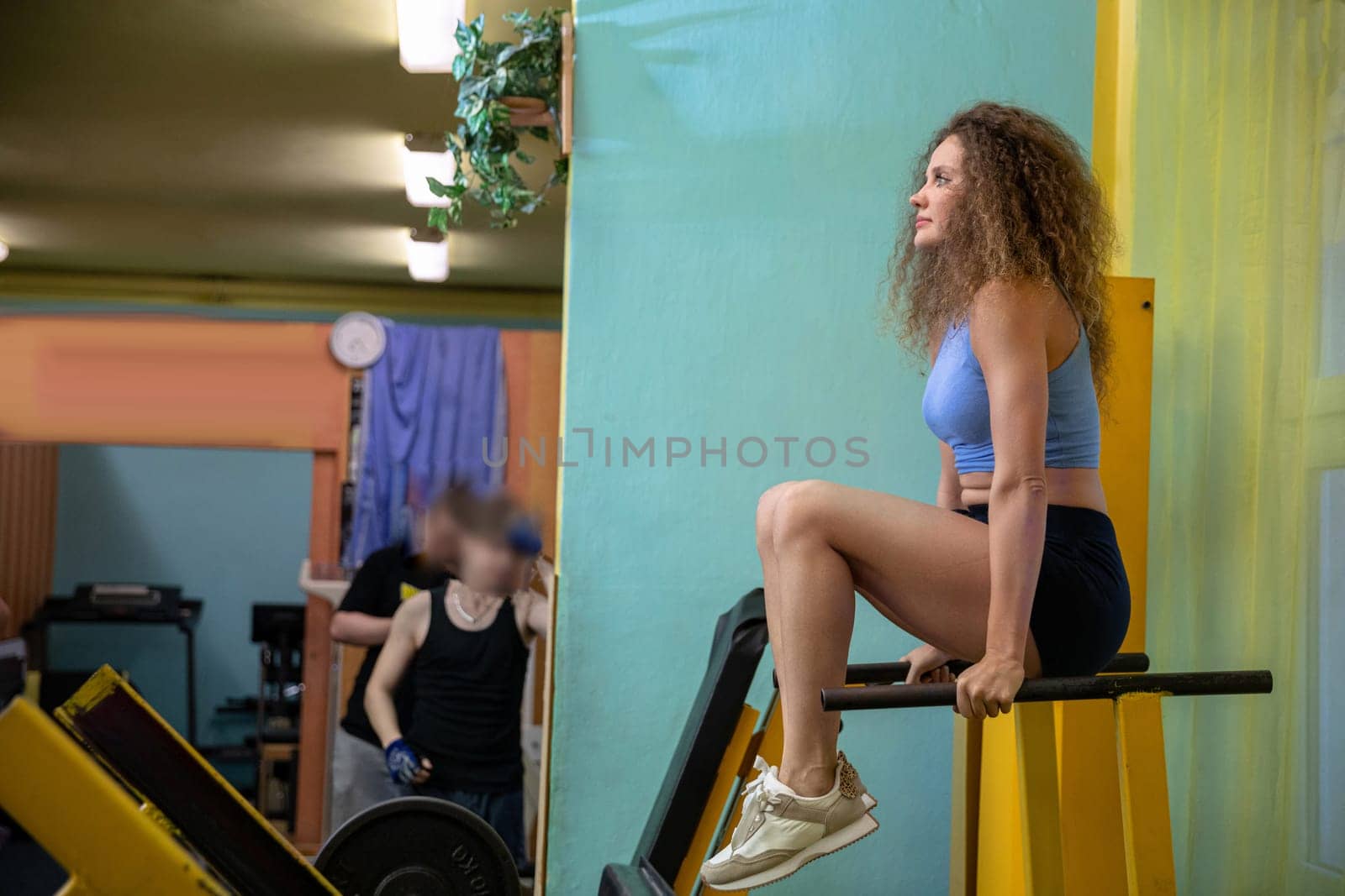 A girl dressed in a tight sports outfit raises herself on her hands. She is holding with her hands two bars on which she is lifting. In the background you can see a mirror hanging on the wall. The silhouettes of several people are reflected in the mirror.