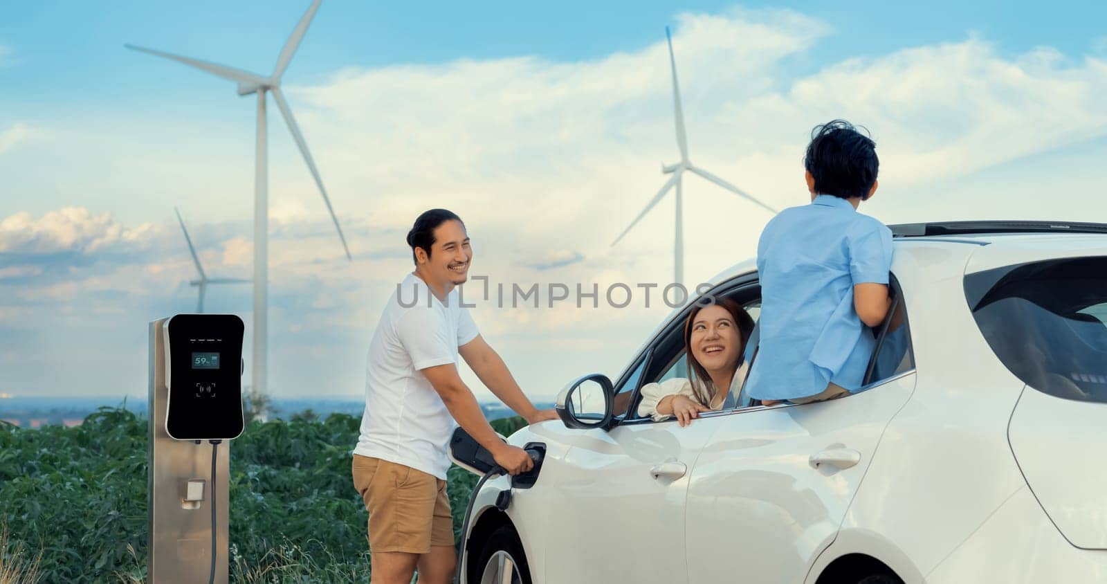 Concept of progressive happy family enjoying their time at wind farm with electric vehicle. Electric vehicle driven by clean renewable energy from wind turbine generator for charging station.