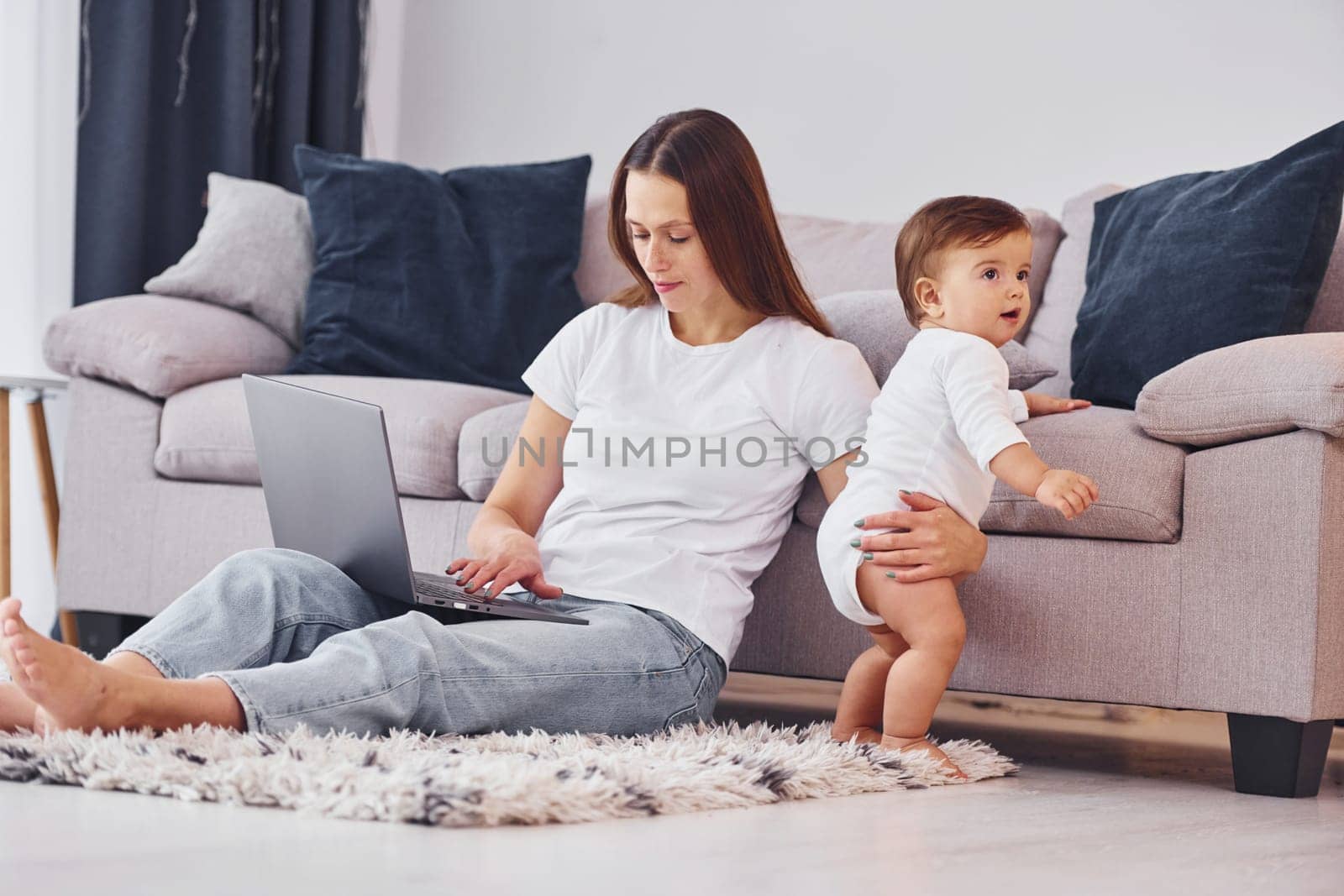 Woman working by using laptop. Mother with her little daughter is indoors at home together.