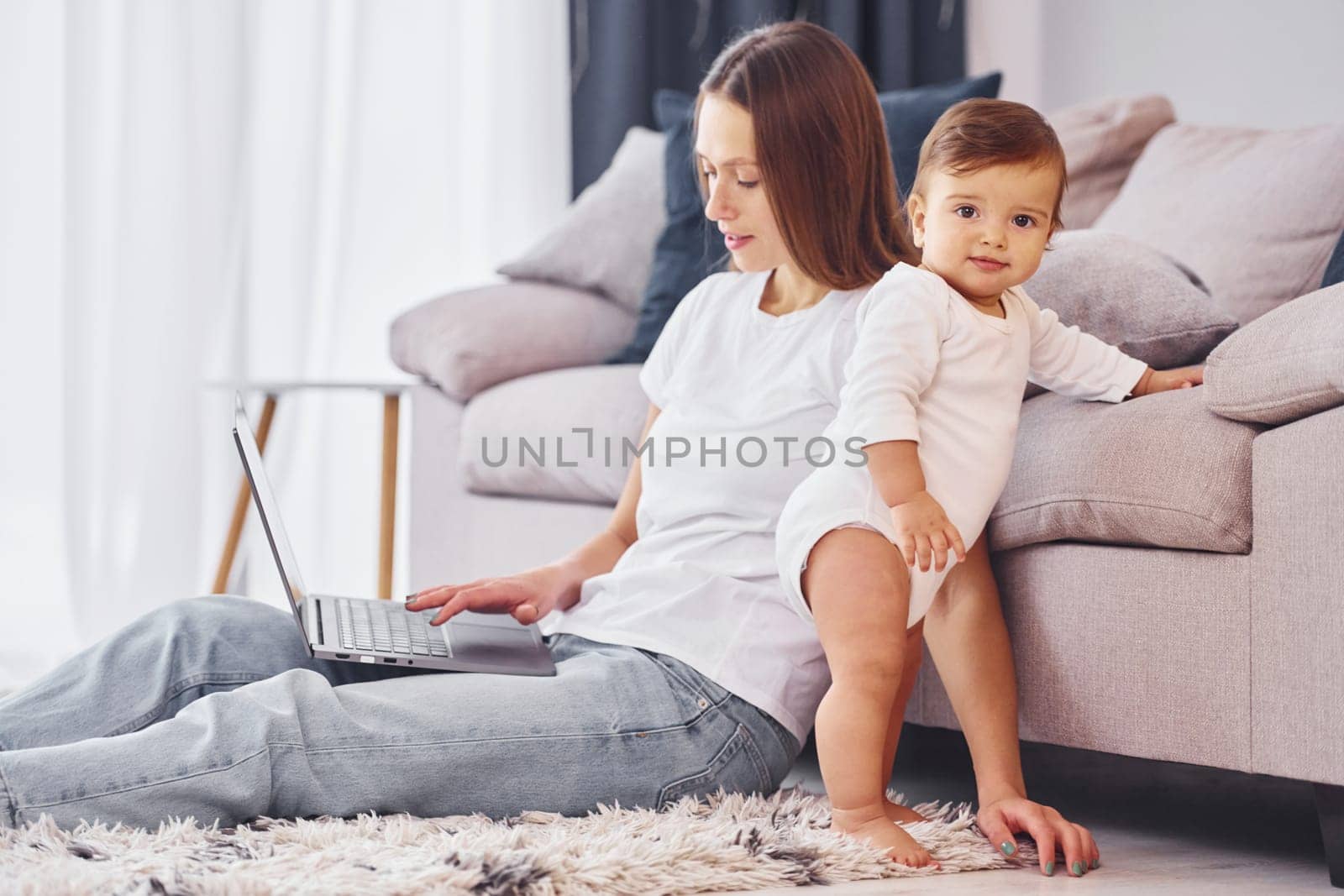 Woman working by using laptop. Mother with her little daughter is indoors at home together.