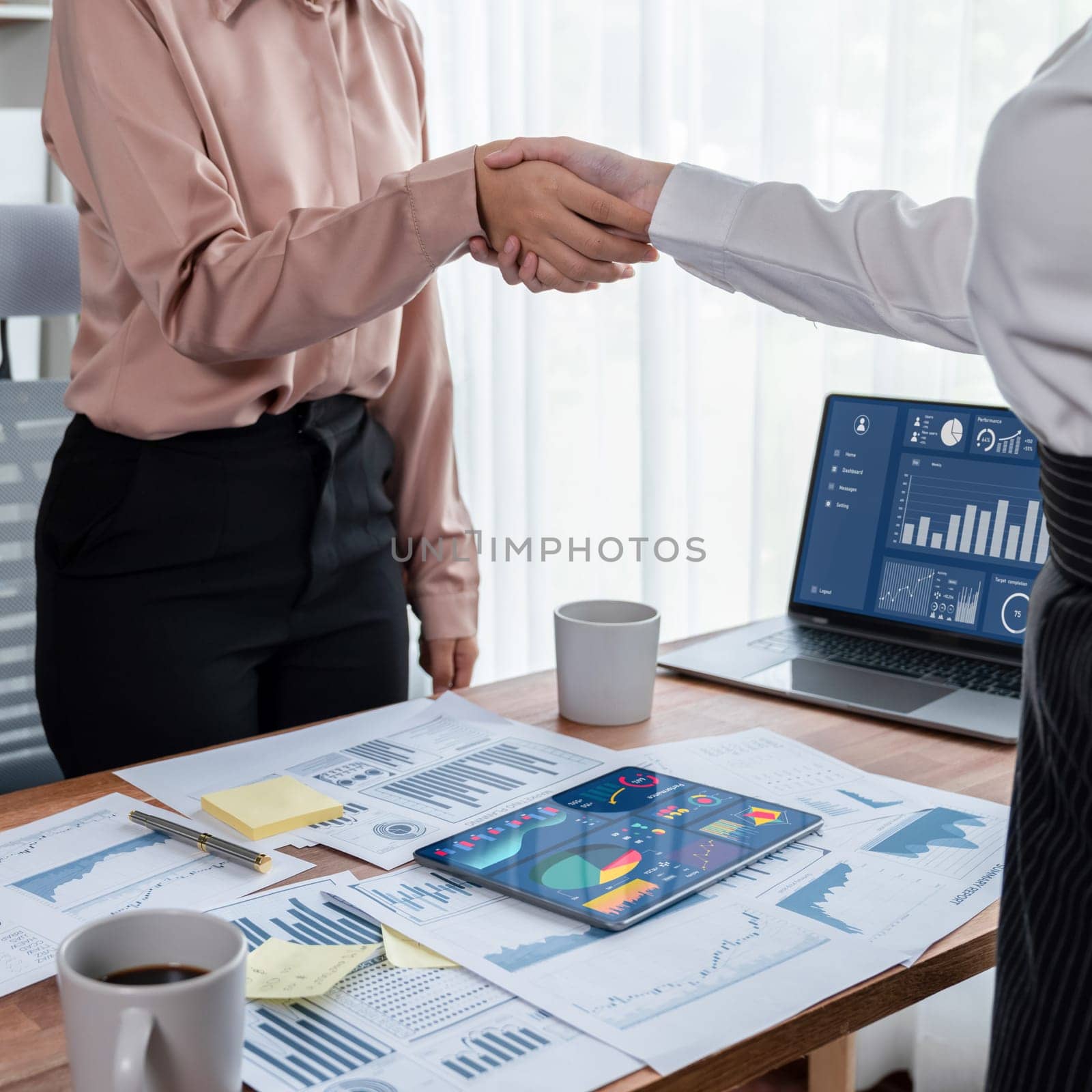 Two Asian businesswomen shaking hands in modern office after successfully analyzing piles of dashboard data papers, as importance of teamwork and integrity in the workplace concept. Enthusiastic