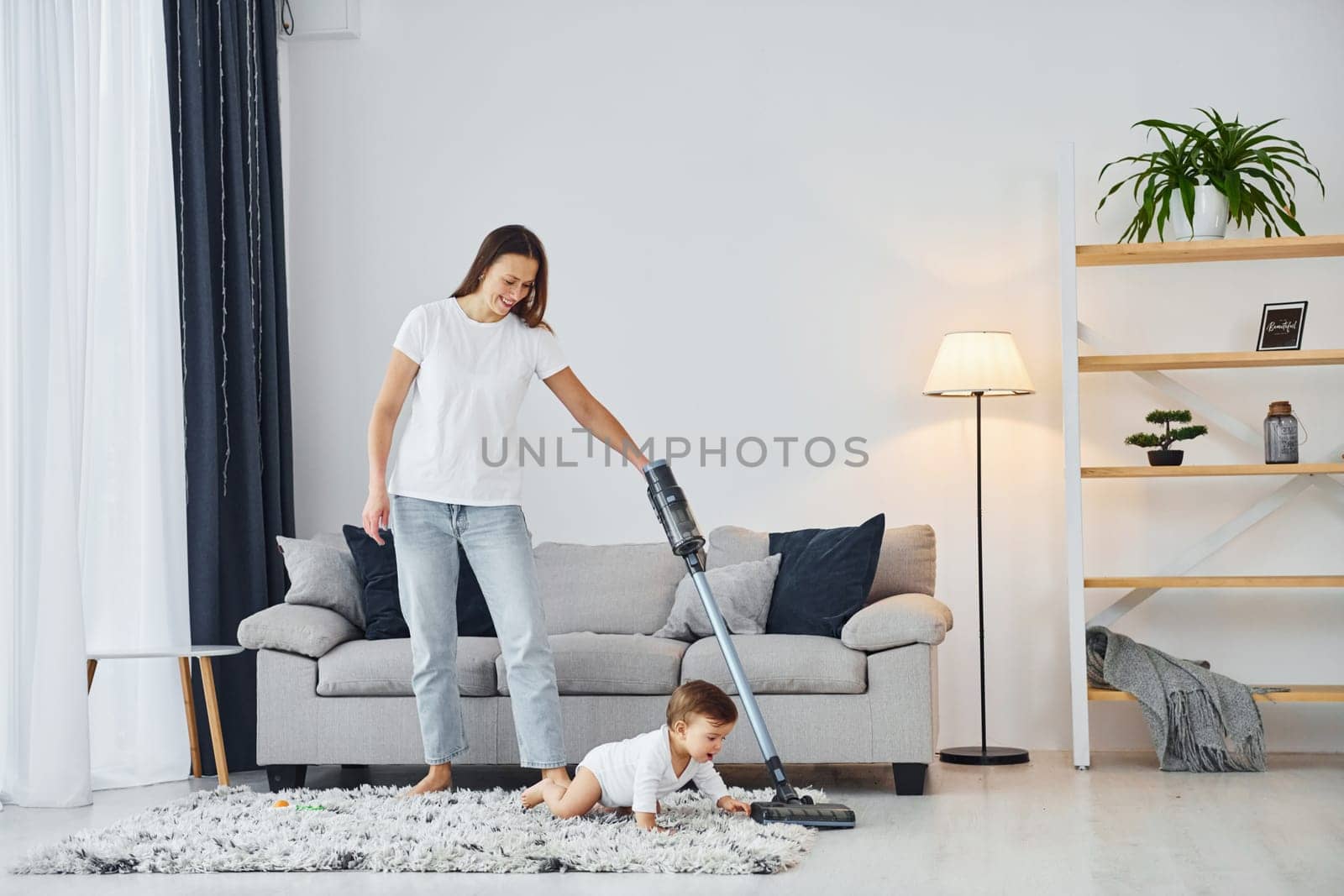 Cleaning floor by using vacuum cleaner. Mother with her little daughter is indoors at home together.