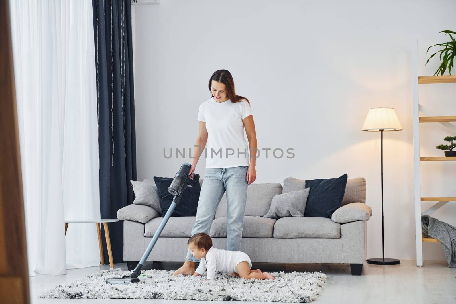 Cleaning floor by using vacuum cleaner. Mother with her little daughter is indoors at home together.