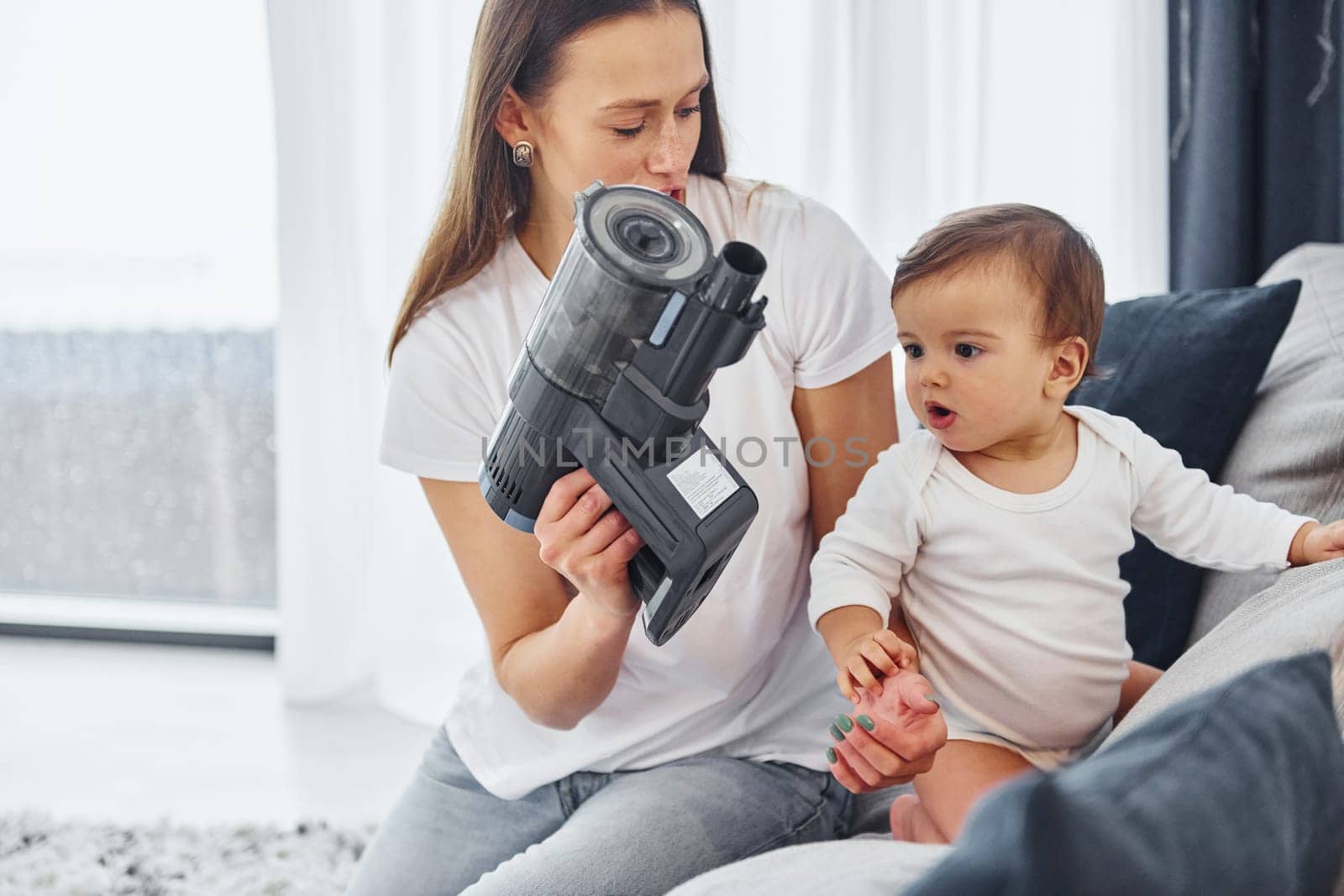 Holding vacuum cleaner. Mother with her little daughter is indoors at home together.