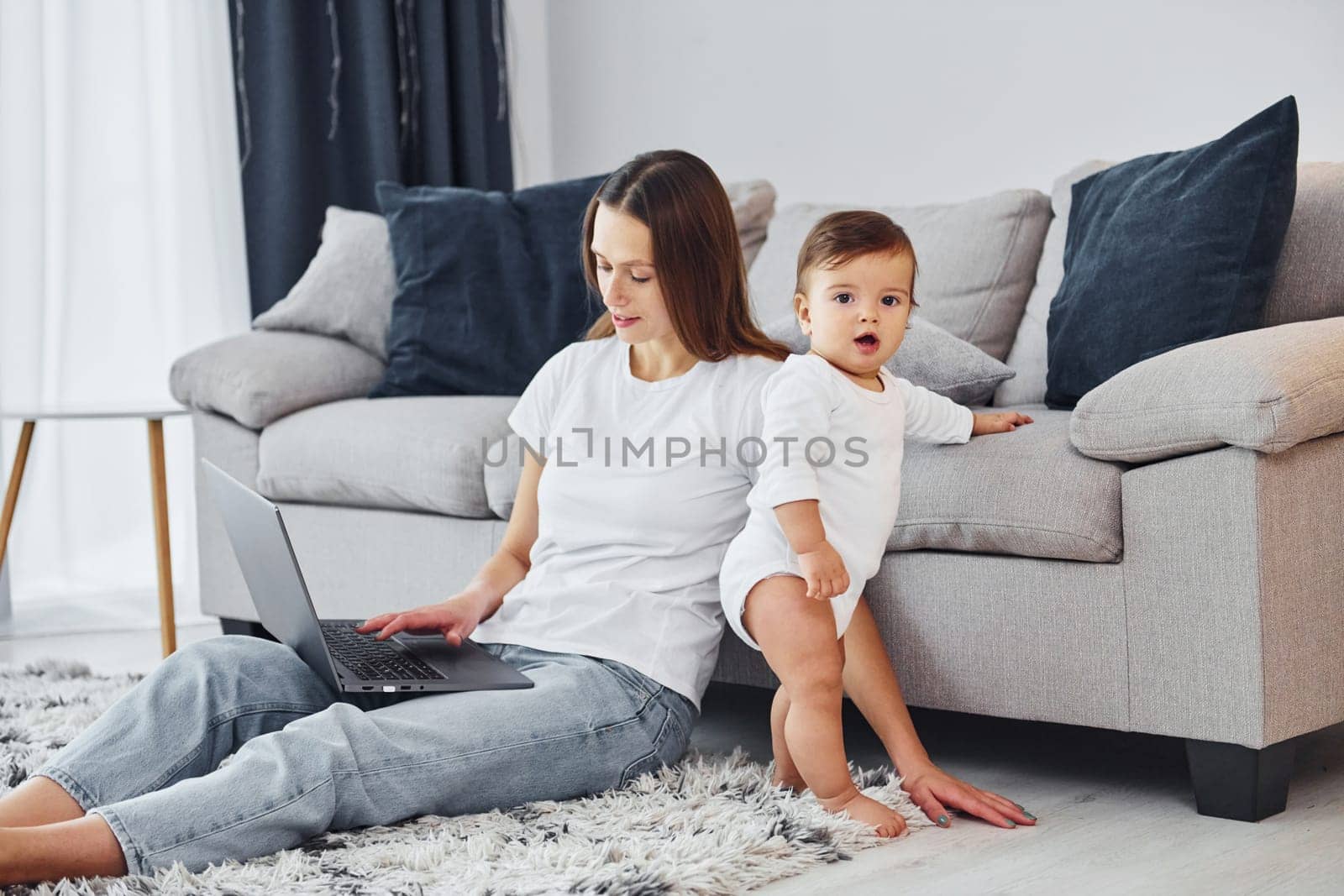 Silver colored laptop. Mother with her little daughter is indoors at home together.