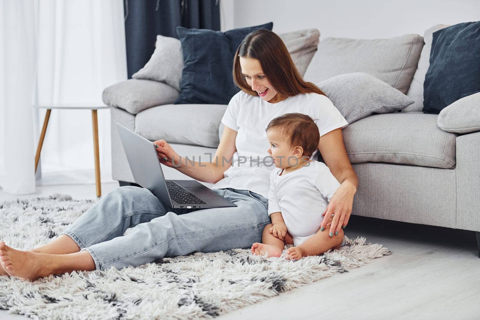 Silver colored laptop. Mother with her little daughter is indoors at home together.