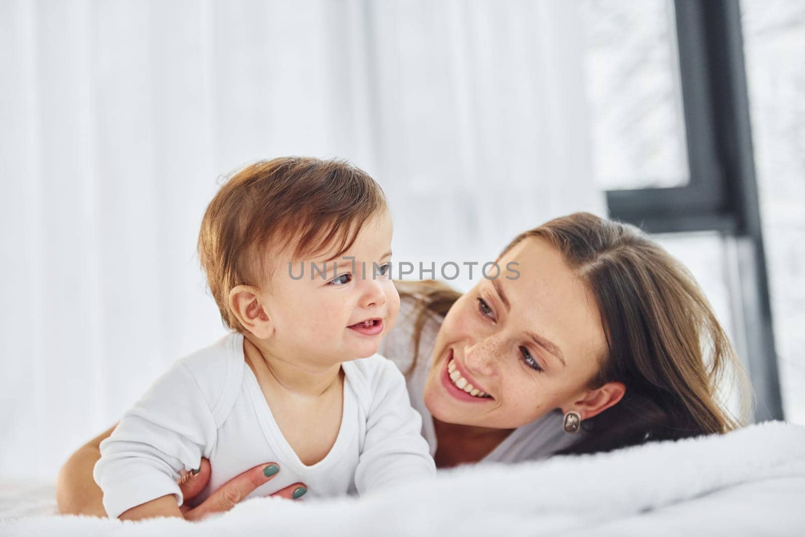 On the white bed. Mother with her little daughter is indoors at home together.