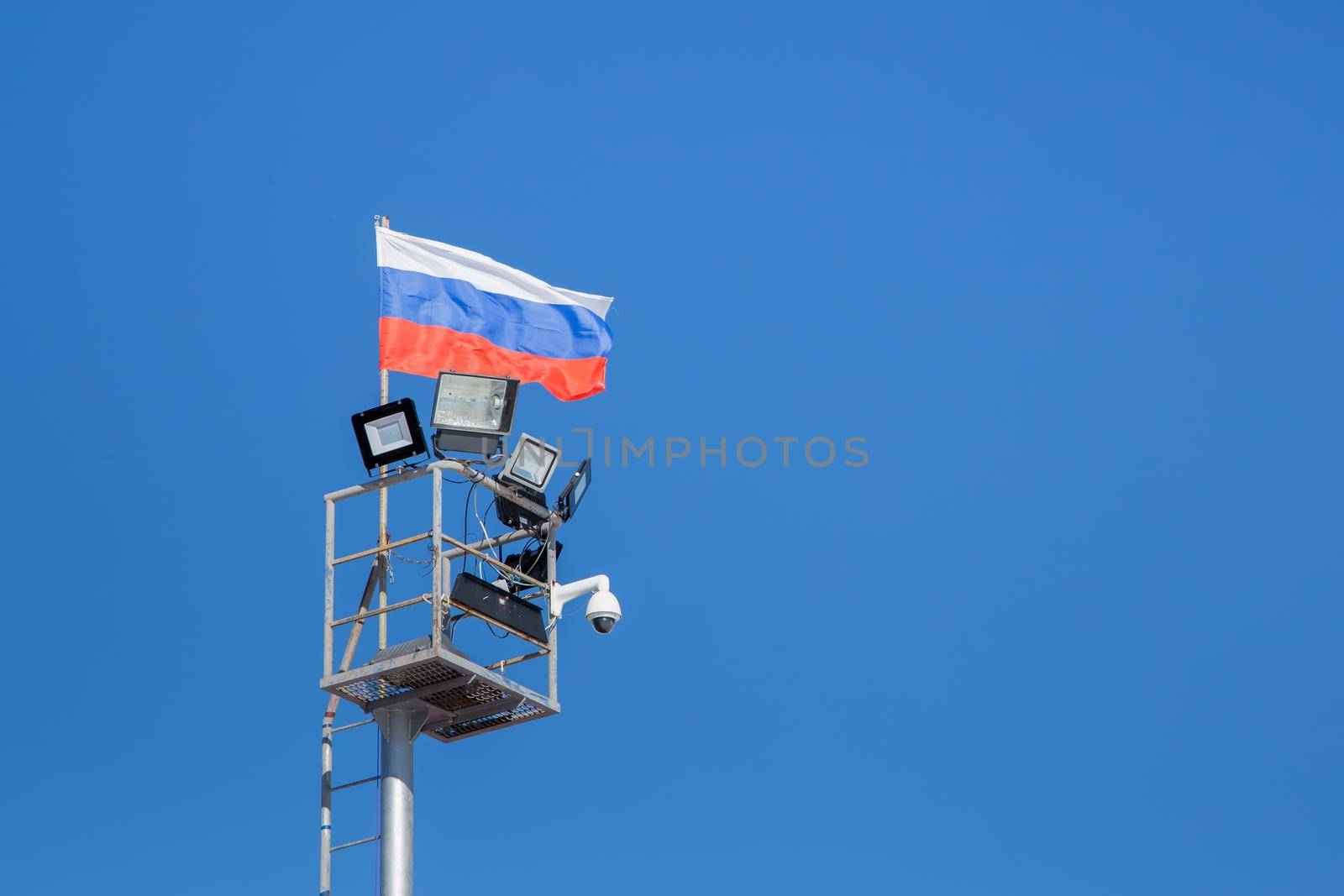 A tower with spotlights and a video surveillance camera. The flag of the Russian Federation develops against a blue sky.