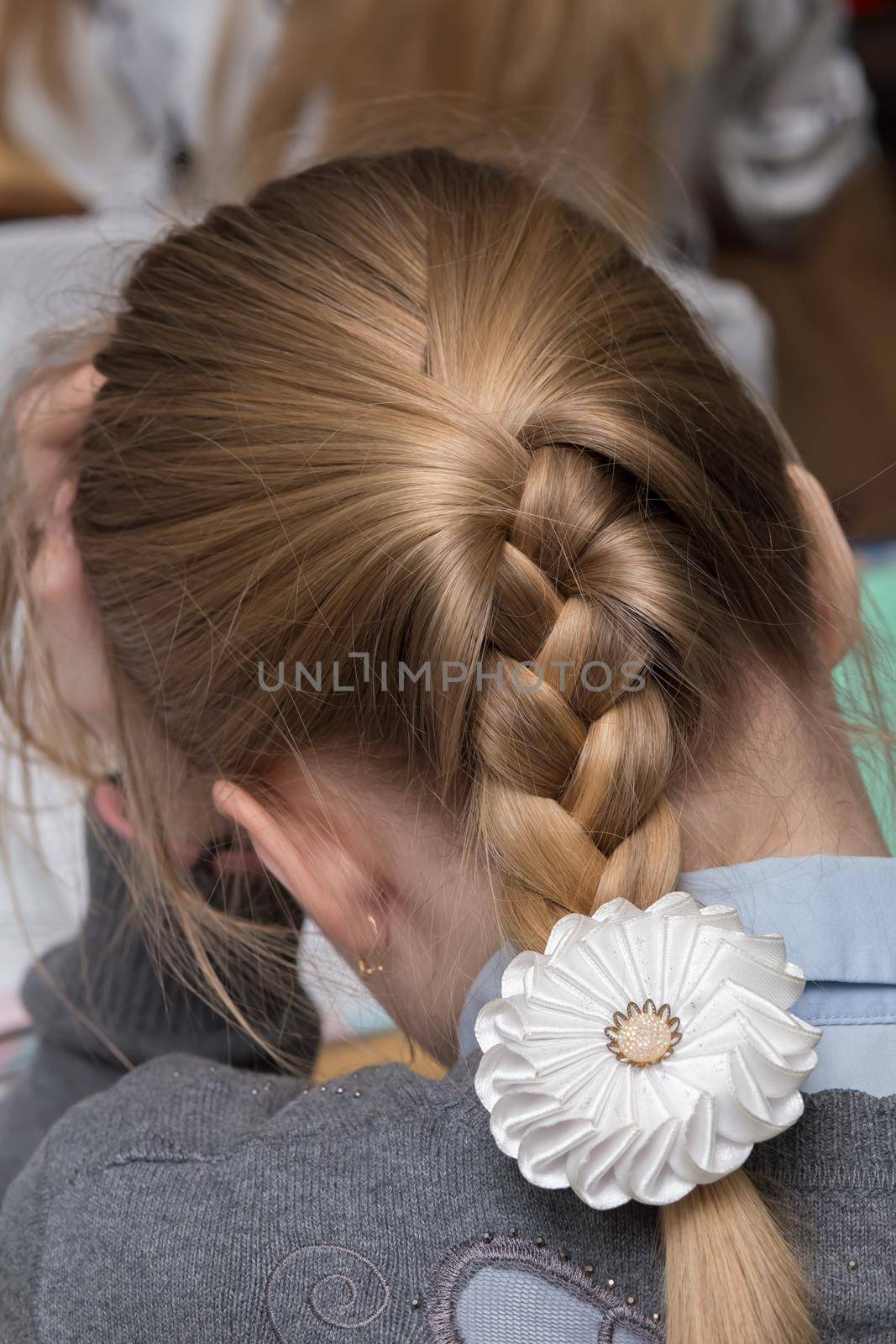 A short pigtail on the back of the girl's head with a bow. The girl rests her head on her hand, she thinks. The student is doing her homework.