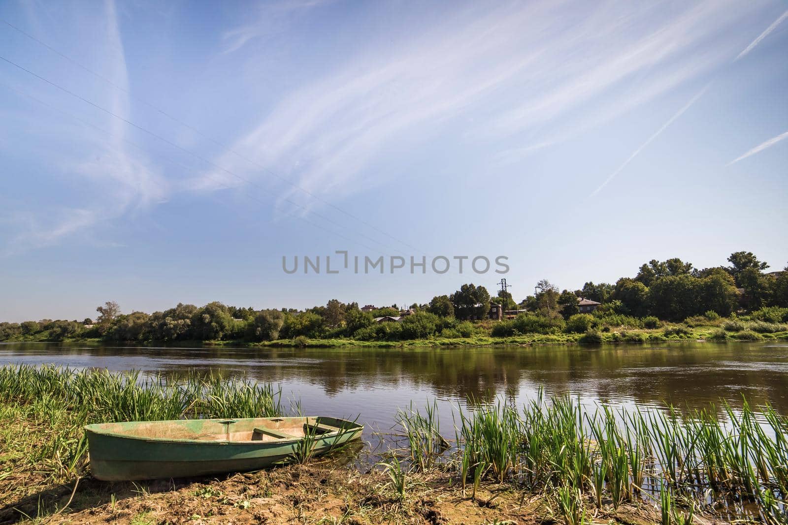 Lonely old boat on the river bank. Tall green grass grows. In the background there is a blue sky and low trees.