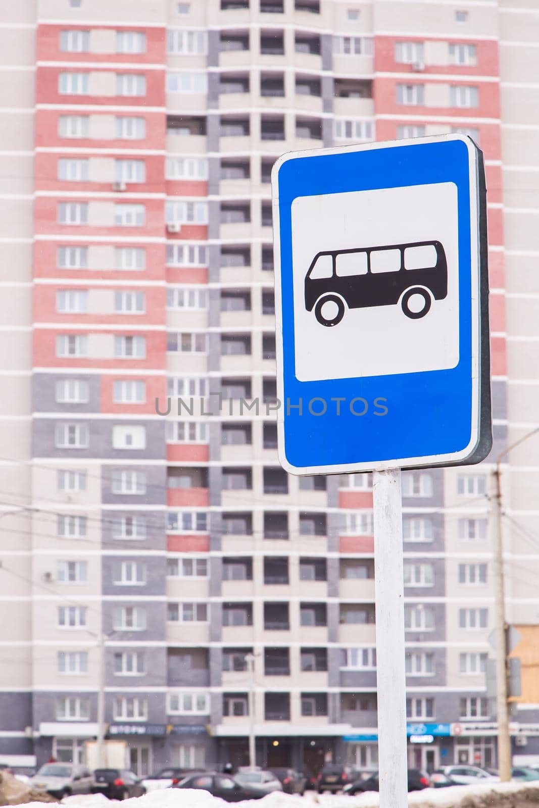 Bus stop road sign on a metal pipe. In the background, a high-rise apartment building is out of focus. Small snowflakes are falling.