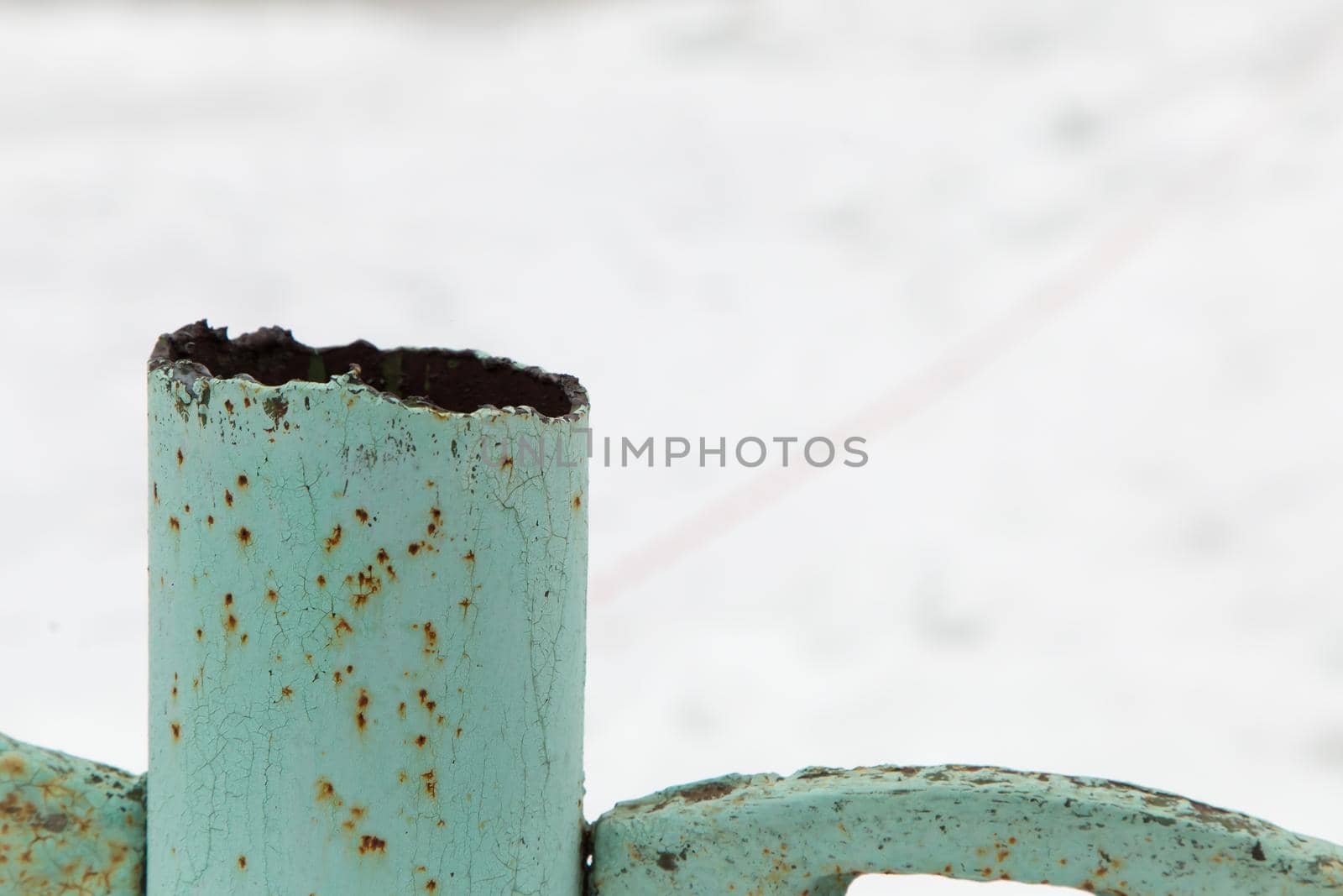 A rusty metal pipe sticks out over an iron fence. The top cut is uneven with very sharp and dangerous edges. Against the background of white snow.