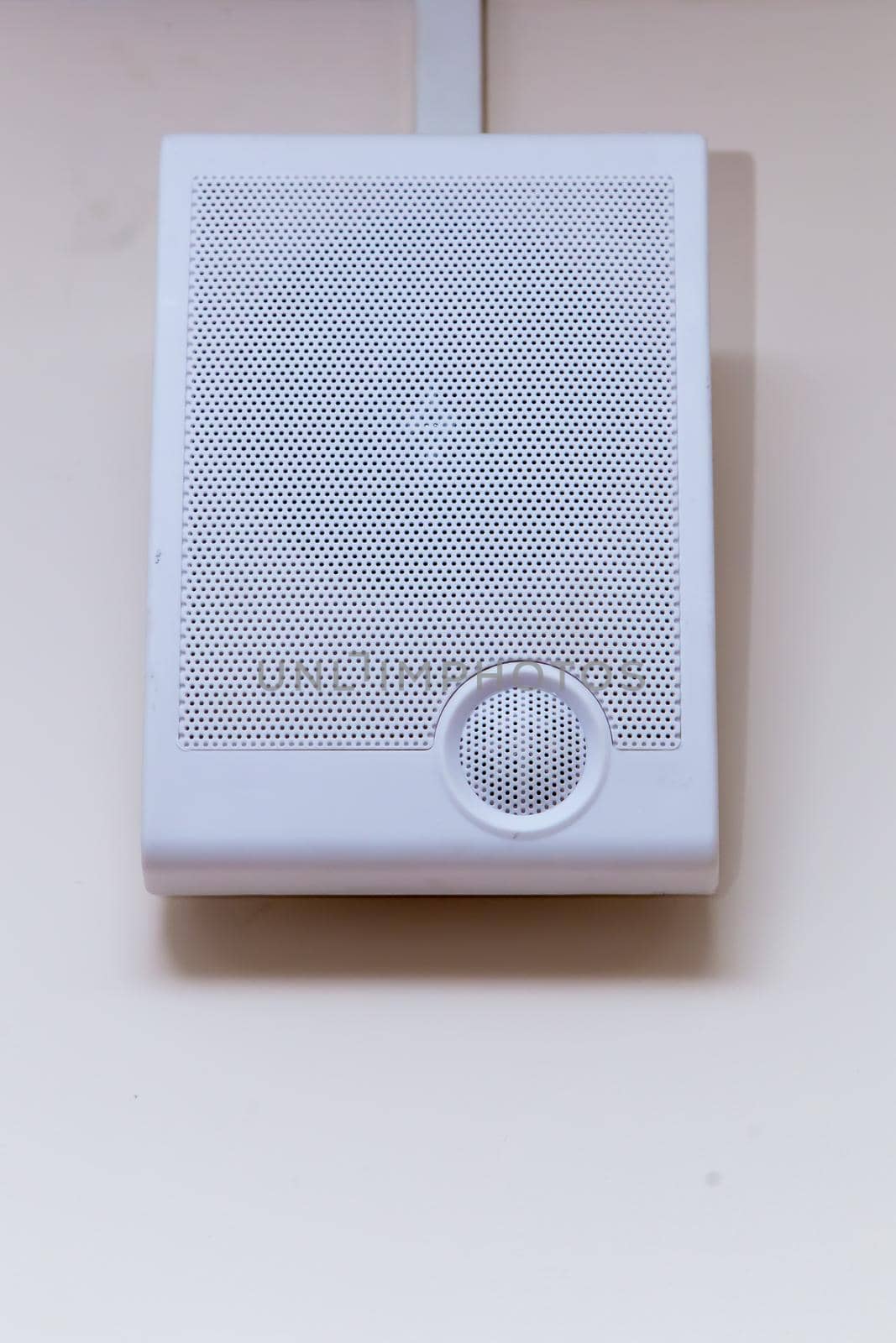 A white radio transmitter hangs on a beige wall. On the front side there is a grill with small holes for a loudspeaker. Electronic device for listening to messages, music and news.