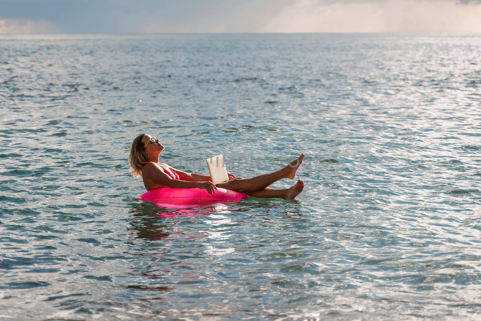 Woman works on laptop in sea. Freelancer, blond woman in sunglases floating on an inflatable big pink donut with a laptop in the sea at sunset. People summer vacation rest lifestyle concept. by Matiunina