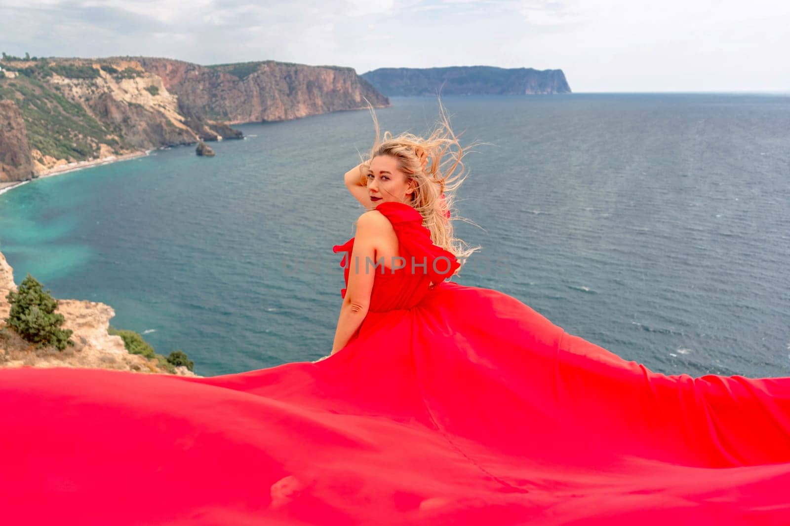 A woman in a red silk dress sits by the ocean with mountains in the background, her dress swaying in the wind