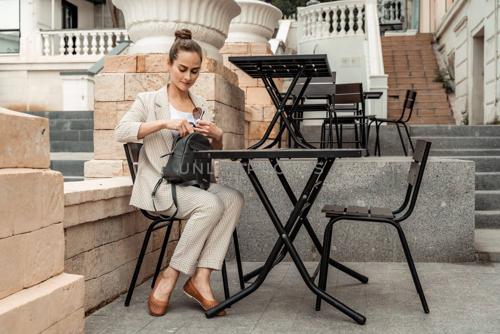 A middle aged woman sits at a table in a cafe in the hands of a black backpack. Dressed in a light suit.