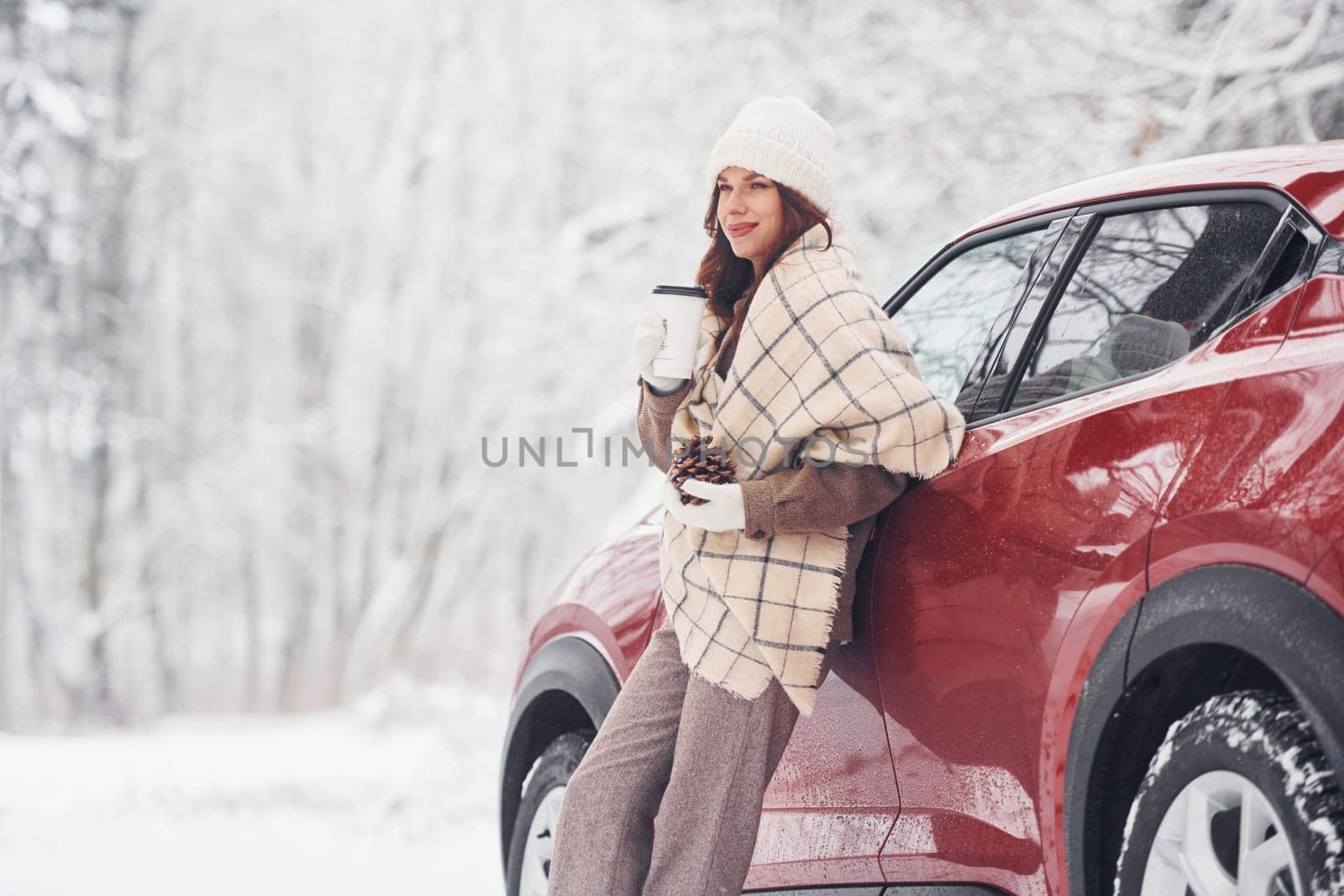 With cup of drink. Beautiful young woman is outdoors near her red automobile at winter time.