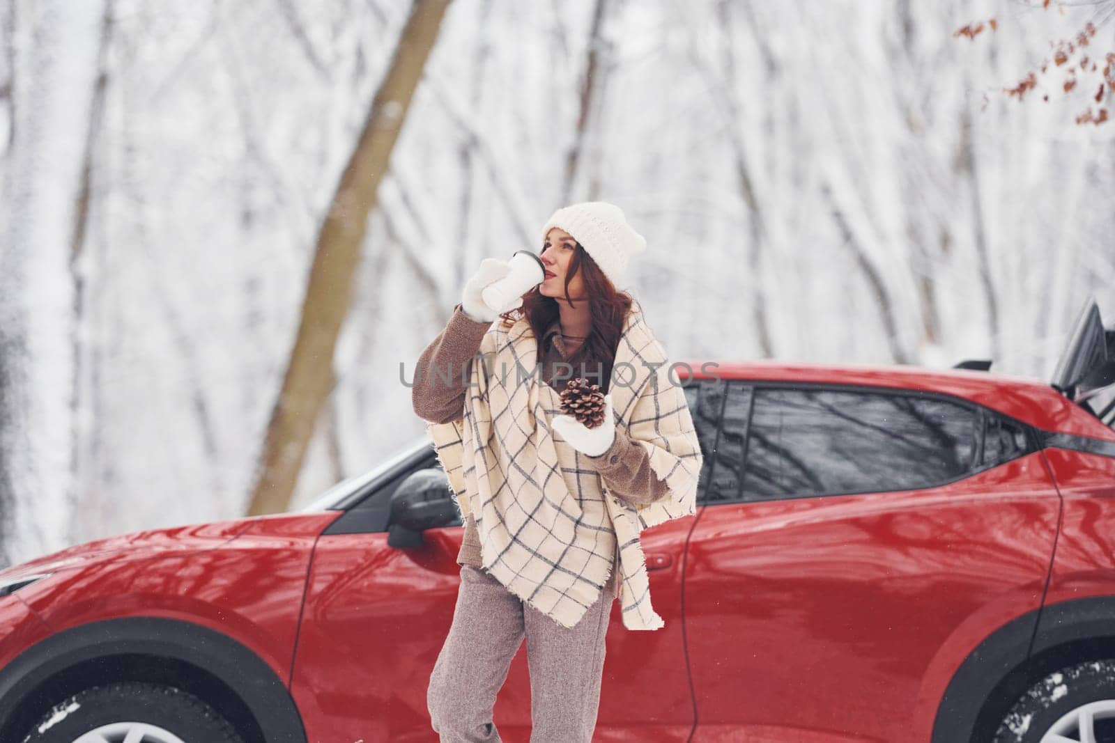 With cup of drink. Beautiful young woman is outdoors near her red automobile at winter time.
