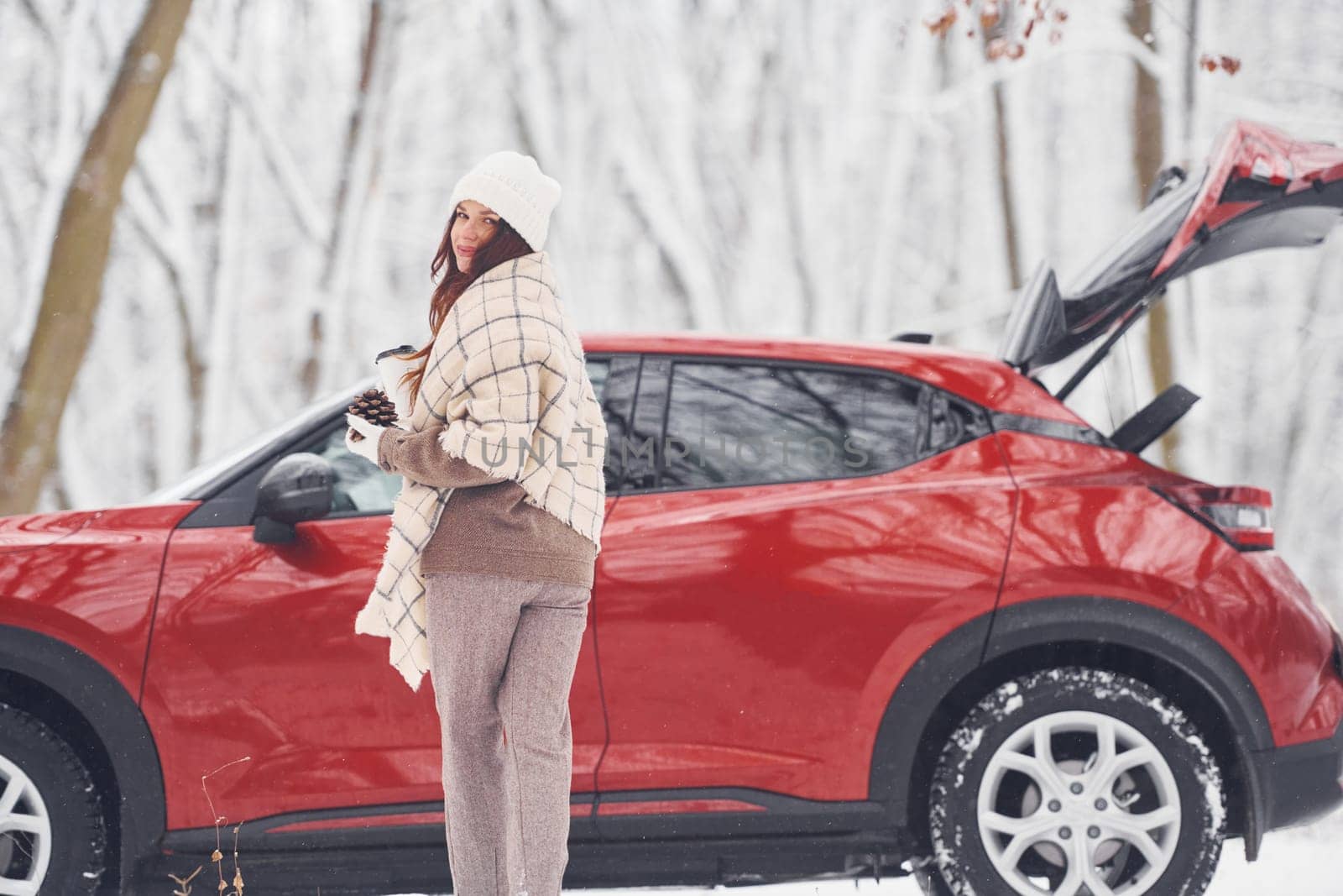 With cup of drink. Beautiful young woman is outdoors near her red automobile at winter time.