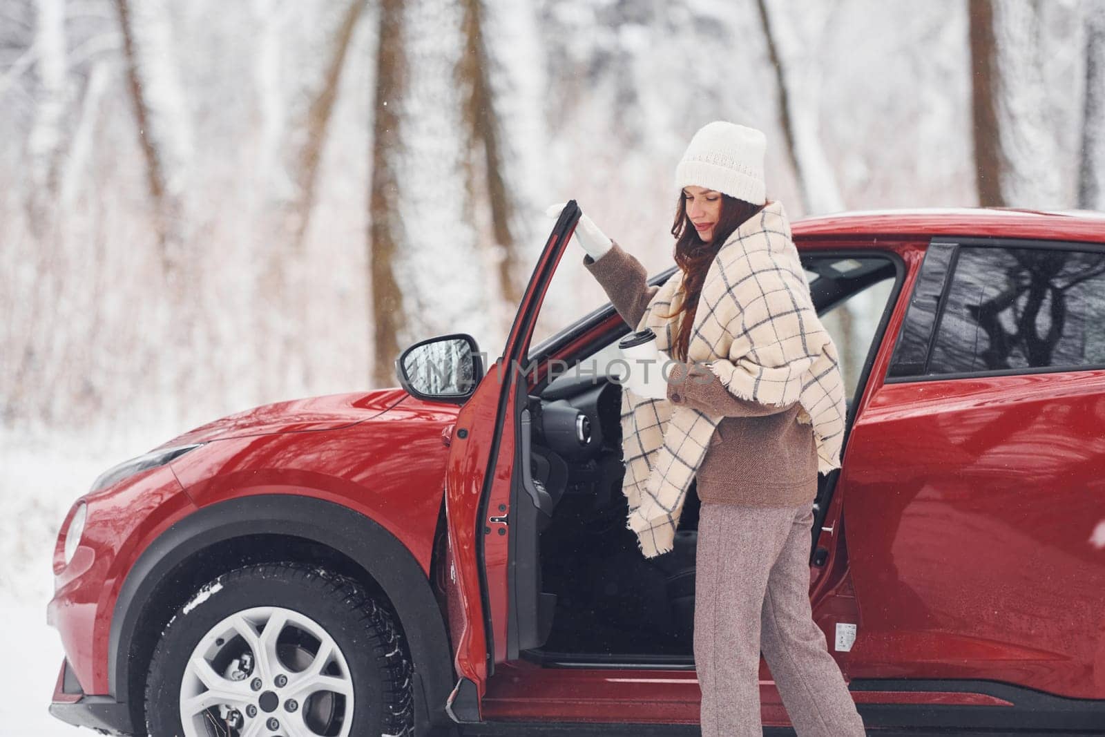 With cup of drink. Beautiful young woman is outdoors near her red automobile at winter time.