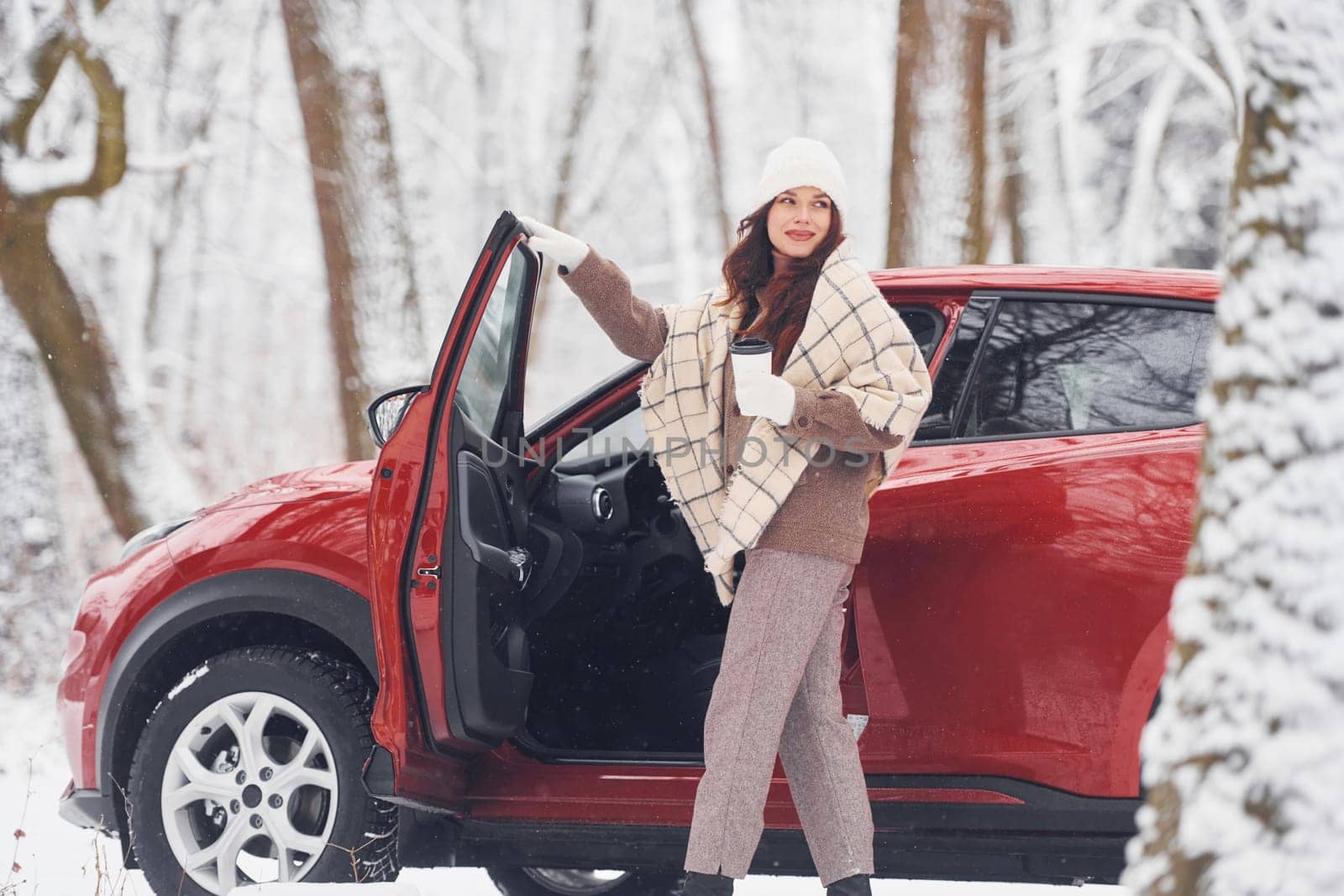 Warm drink in cup. Beautiful young woman is outdoors near her red automobile at winter time.