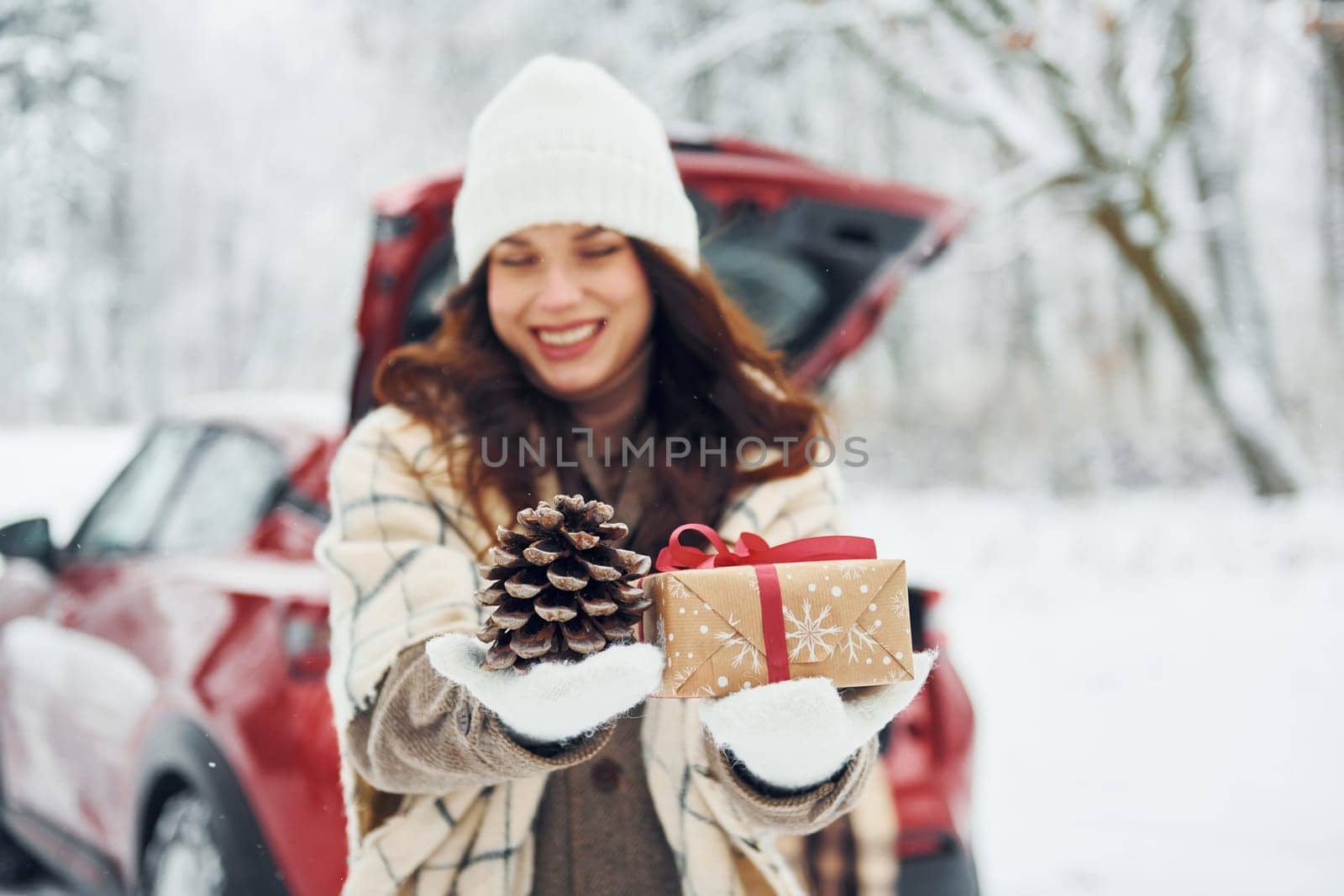 Holding gift box. Beautiful young woman is outdoors near her red automobile at winter time.