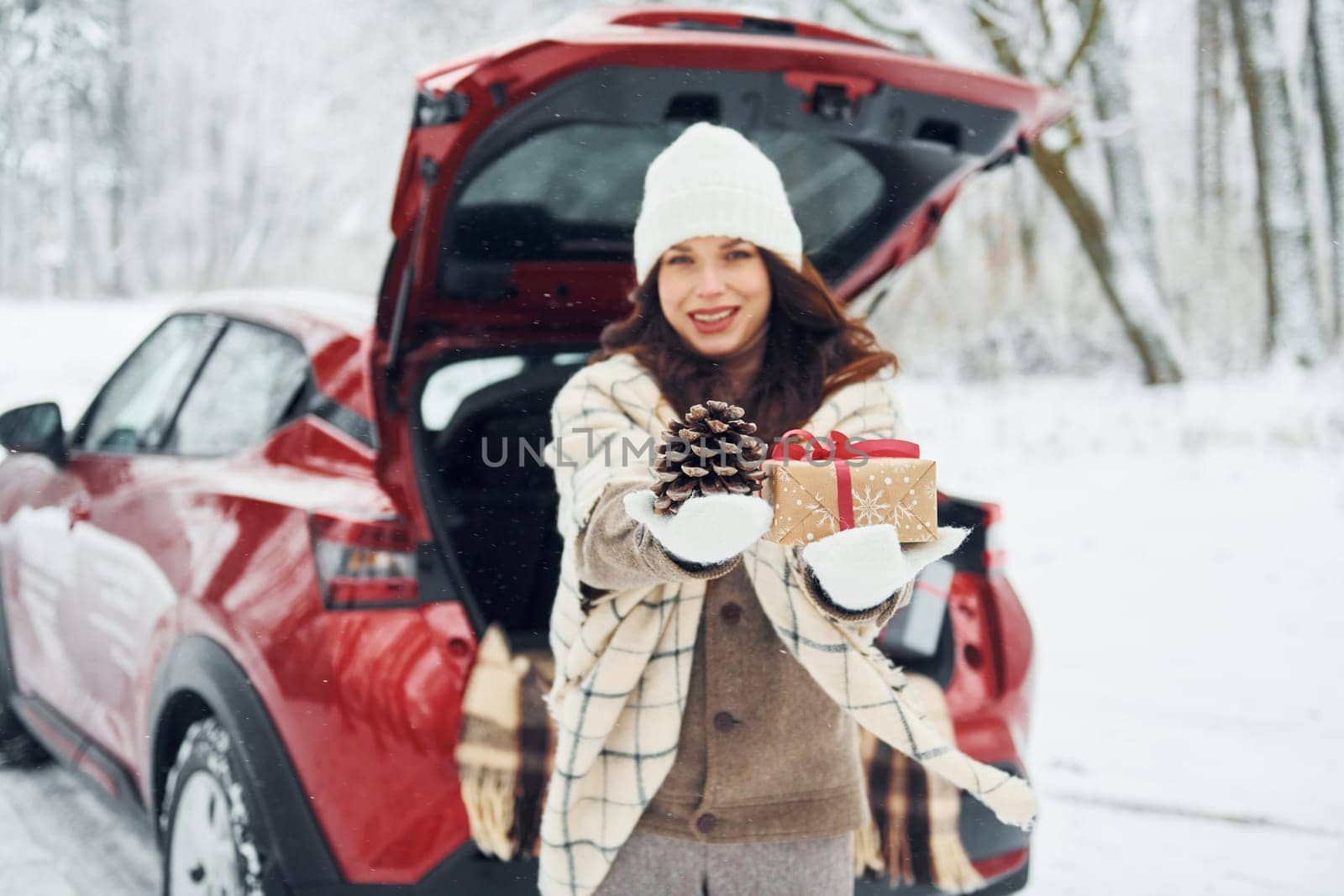 Holding gift box. Beautiful young woman is outdoors near her red automobile at winter time.