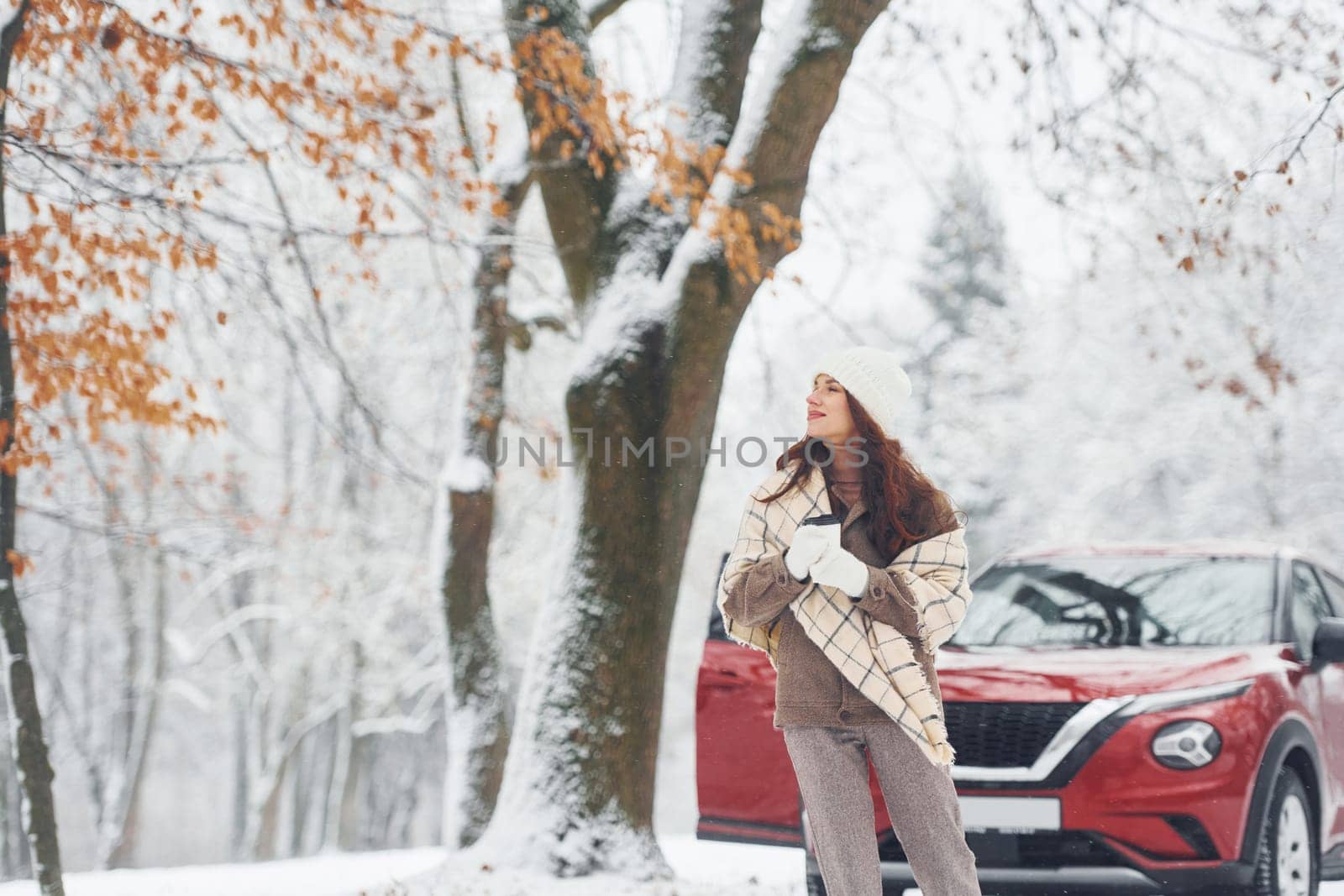 In the forest. Beautiful young woman is outdoors near her red automobile at winter time.