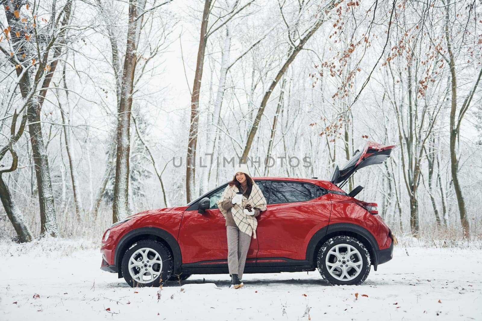 In the forest. Beautiful young woman is outdoors near her red automobile at winter time.