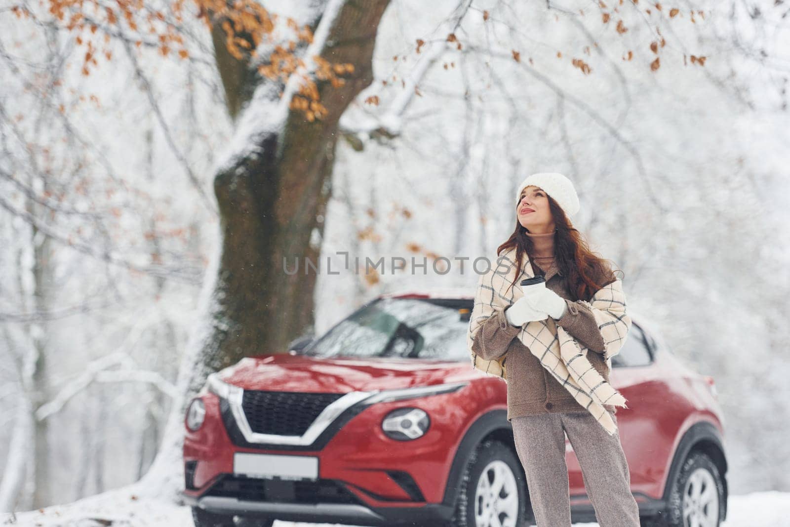 Rural scene. Beautiful young woman is outdoors near her red automobile at winter time.