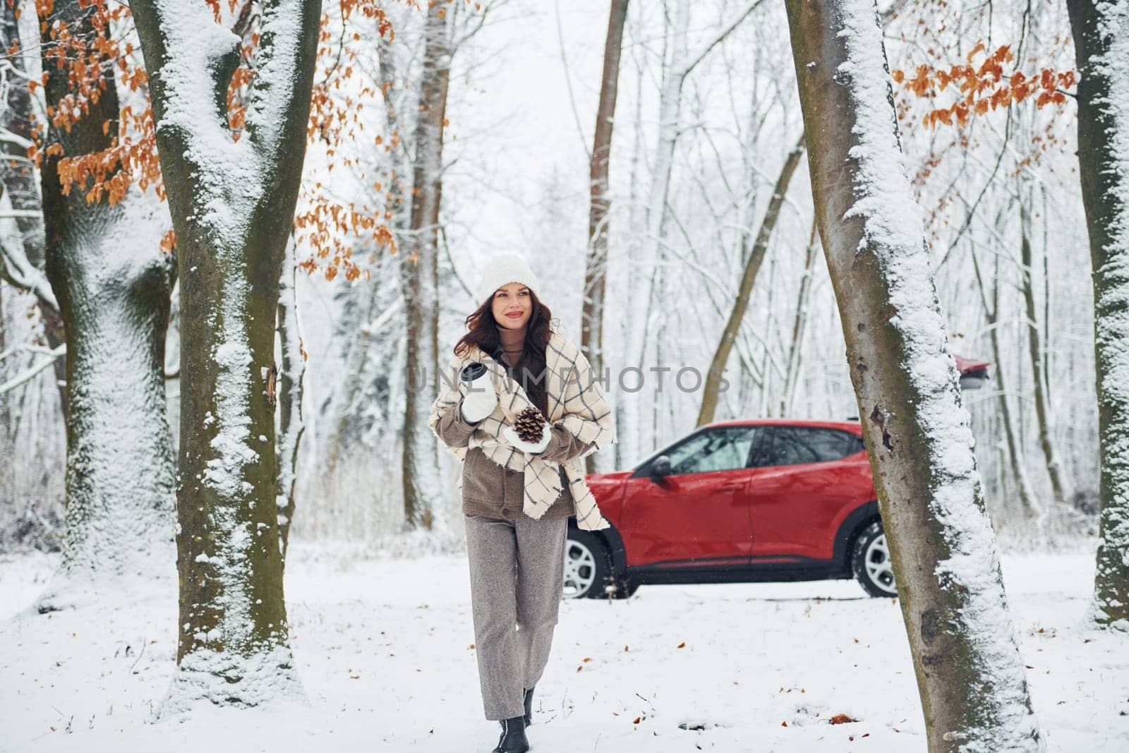 Trees are covered with snow. Beautiful young woman is outdoors near her red automobile at winter time.