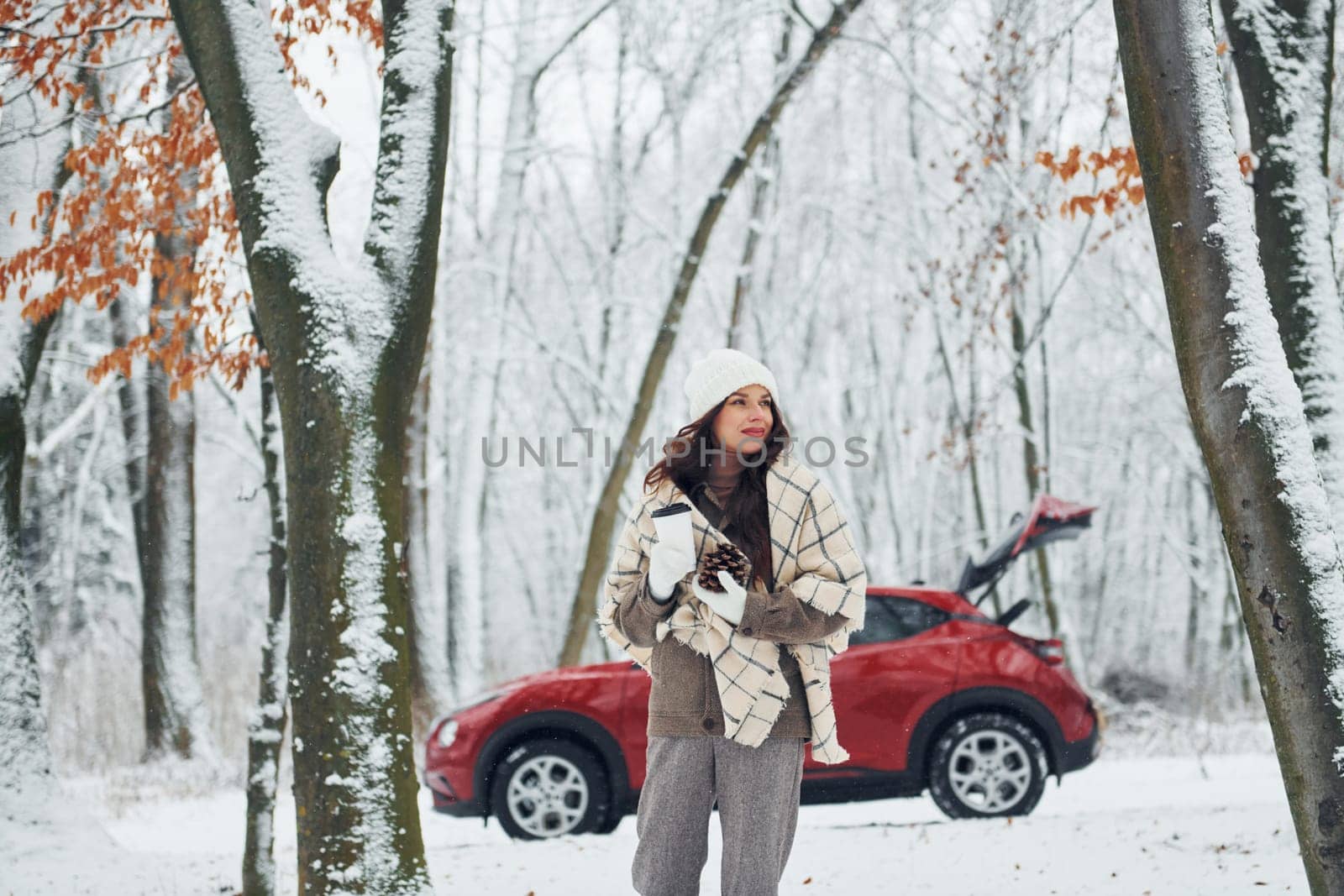 Having a walk in the forest. Beautiful young woman is outdoors near her red automobile at winter time.