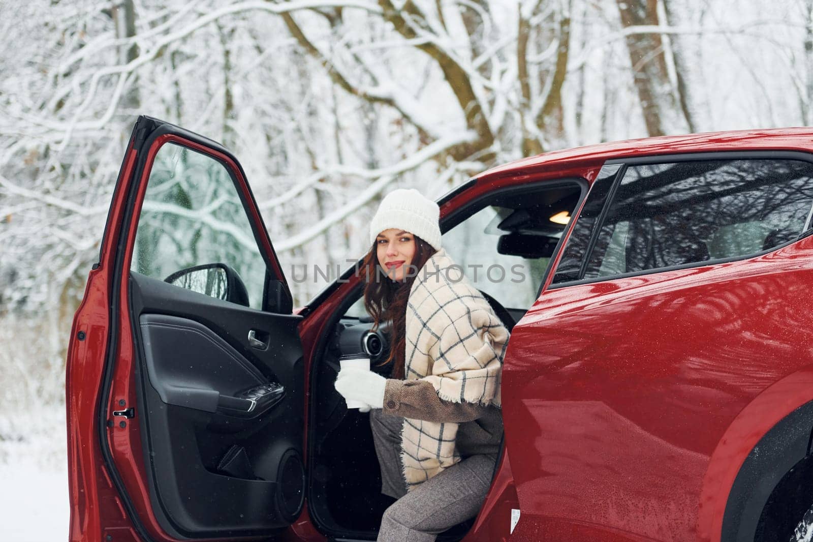 New car is in the forest. Beautiful young woman is outdoors near her red automobile at winter time.