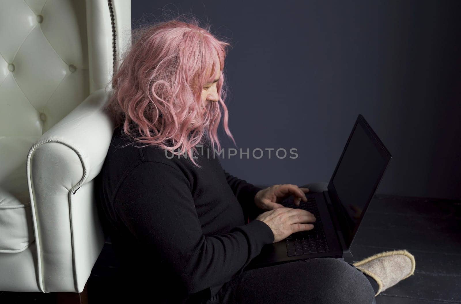 A man in a pink wig sits on the floor near a white chair and works at a computer. Side view